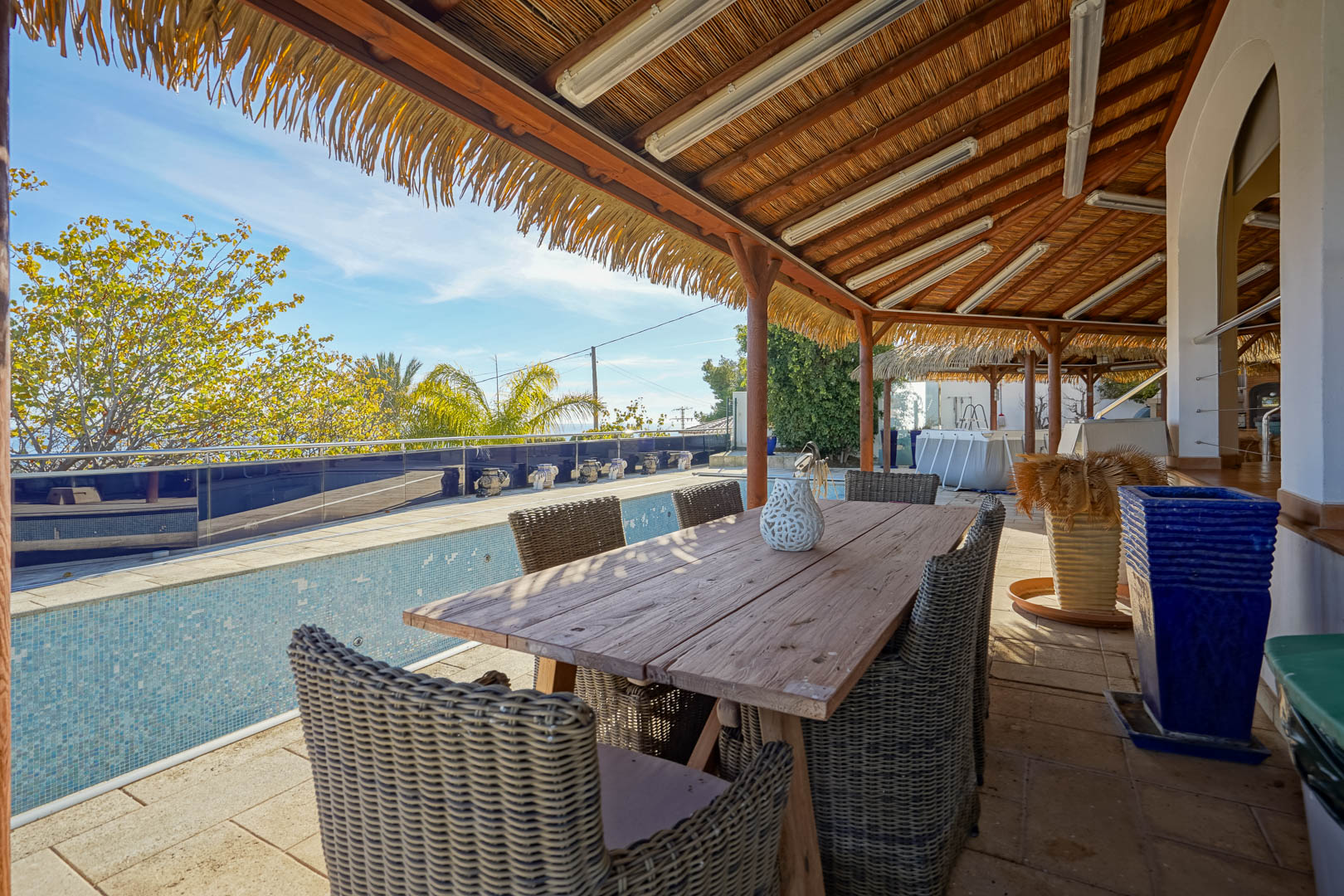 Terraza cubierta con pérgola de brezo, mesa de comedor de madera rústica y sillas de mimbre junto a la piscina con vistas despejadas y entorno ajardinado.