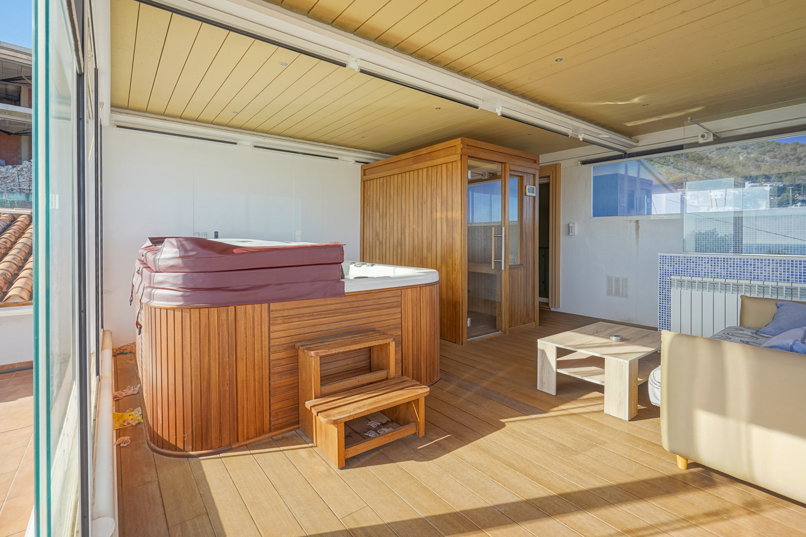 Terraza superior acristalada con zona de bienestar, que incluye un jacuzzi revestido en madera, sauna finlandesa y suelos de tarima bajo un techo técnico con iluminación integrada.
