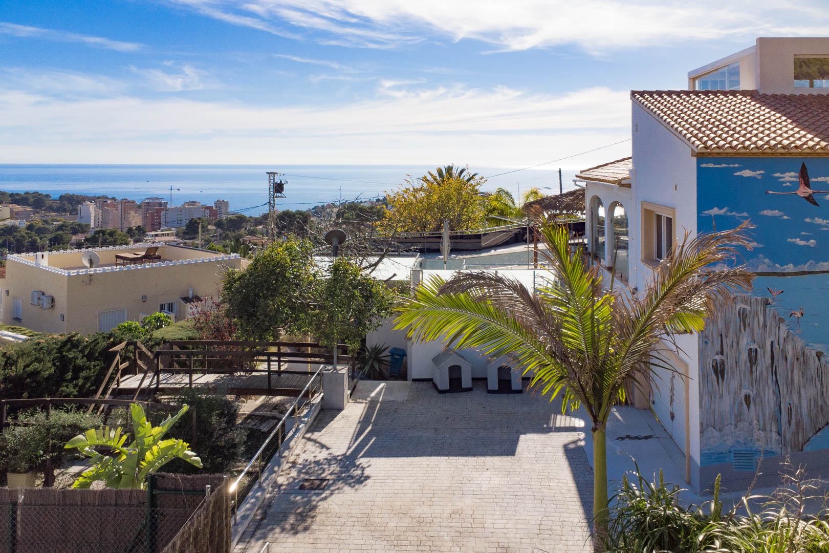 Vista exterior con entrada pavimentada, mural de flamencos en la fachada y vistas panorámicas al mar Mediterráneo y al skyline de Calpe.