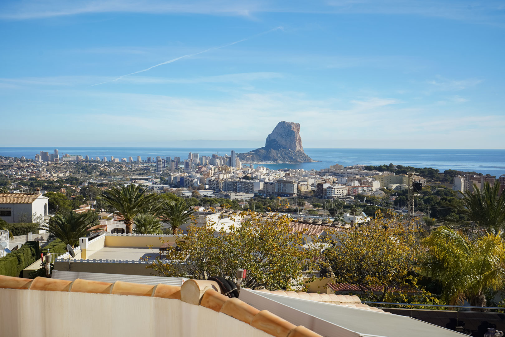 Vistas panorámicas desde la terraza hacia el Peñón de Ifach, el skyline de Calpe y el mar Mediterráneo, enmarcadas por la vegetación local y tejados tradicionales.