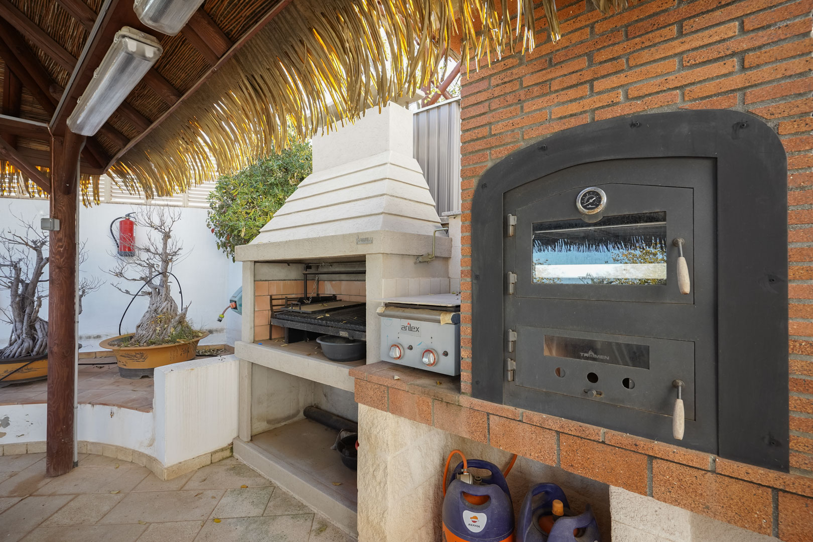 Zona de cocina exterior con horno Tromen, barbacoa de obra y plancha de gas, protegida por una pérgola de madera con cubierta de brezo natural.
