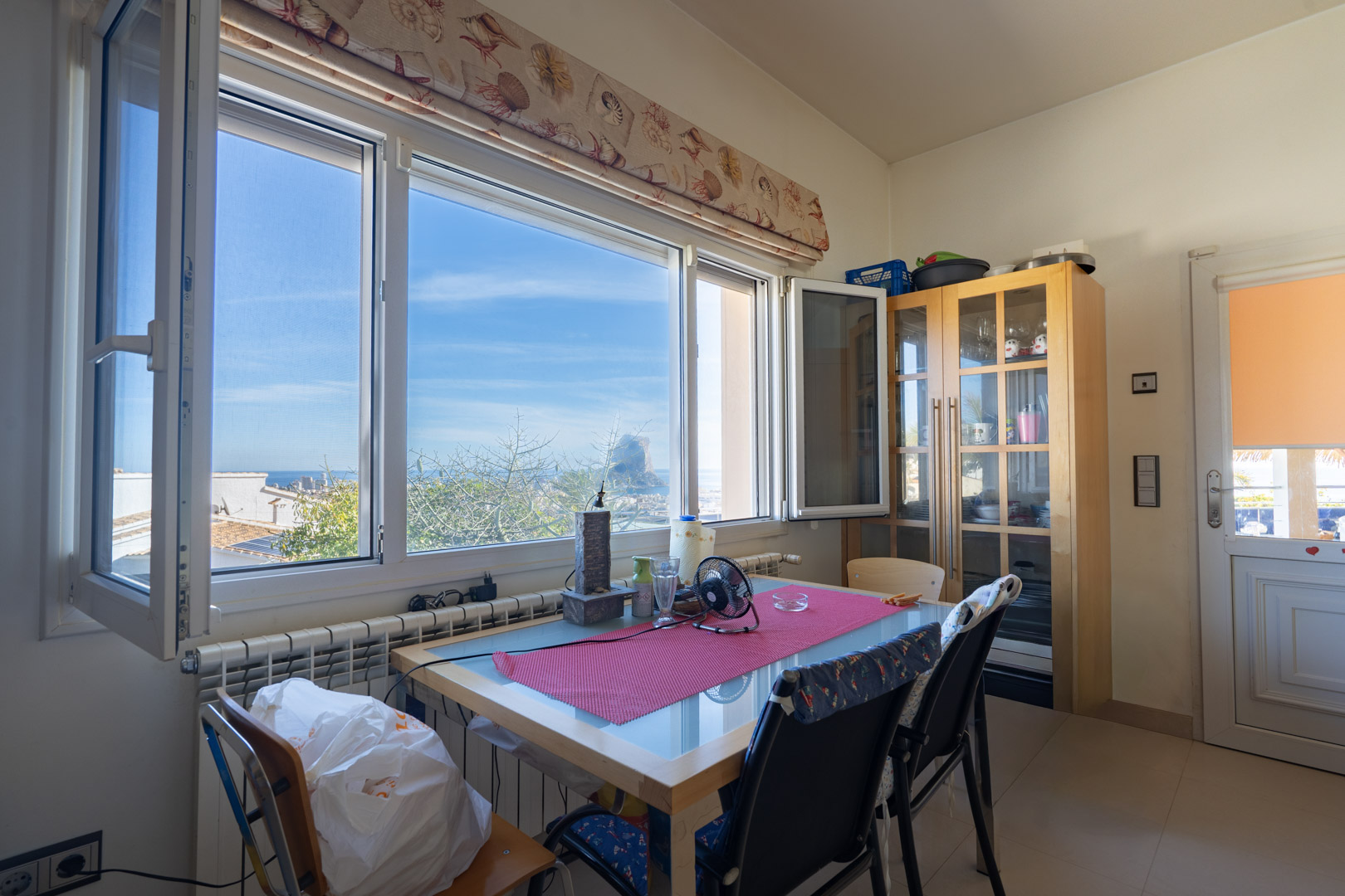 Zona de comedor con vistas directas al Peñón de Ifach. Cuenta con un amplio ventanal, mesa de madera con sobre de cristal y una vitrina clásica de almacenamiento.