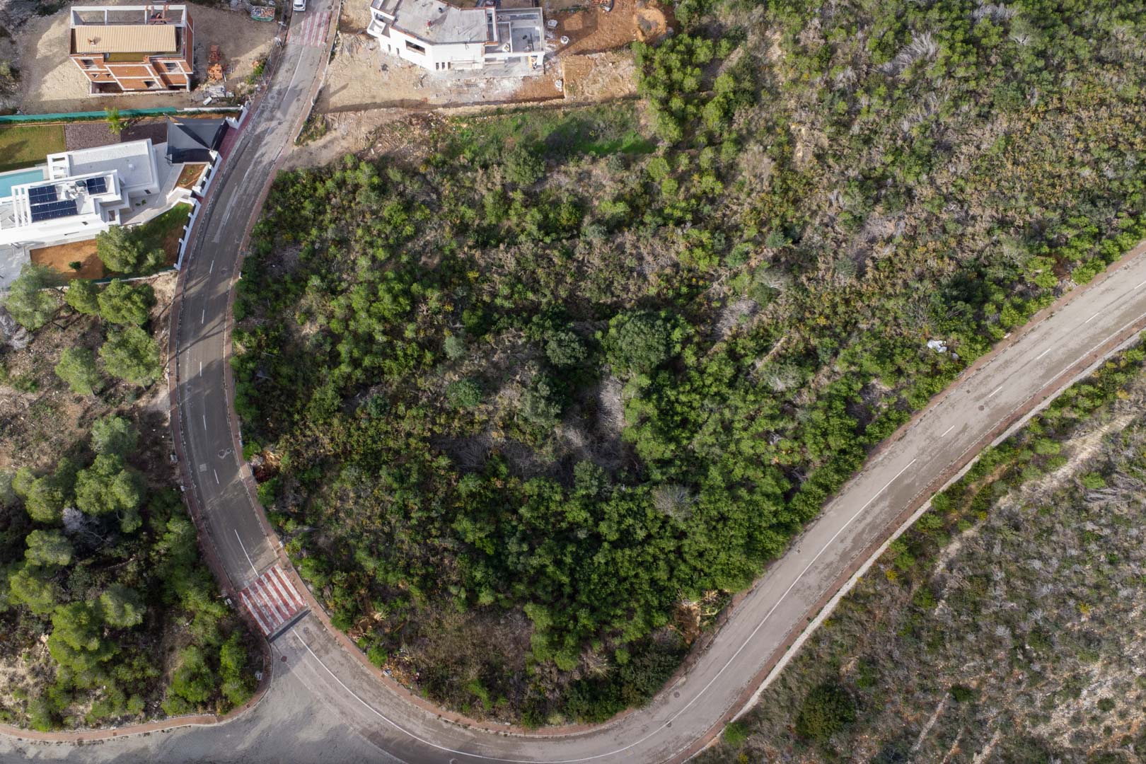 Vista aérea cenital de la parcela urbana en esquina, rodeada de calles pavimentadas con aceras y vegetación mediterránea autóctona en Monte Olímpico.
