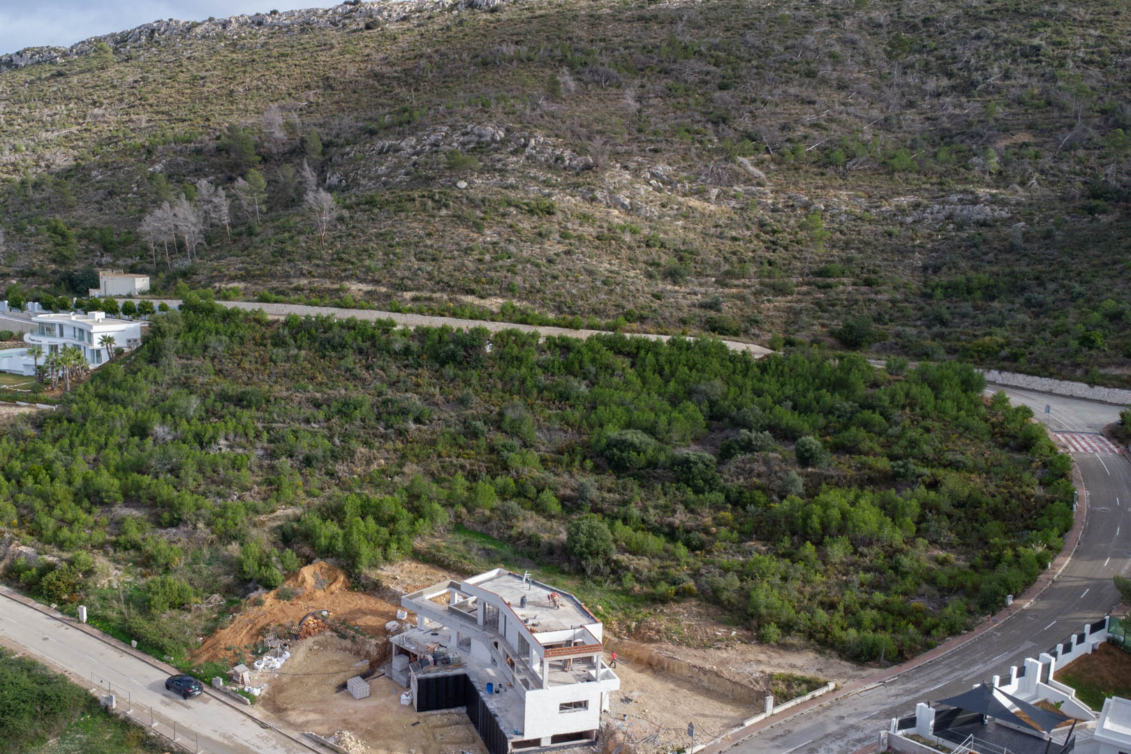 Vista aérea de la parcela en Monte Olímpico, mostrando la infraestructura urbana completada y una villa en construcción rodeada de vegetación mediterránea.
