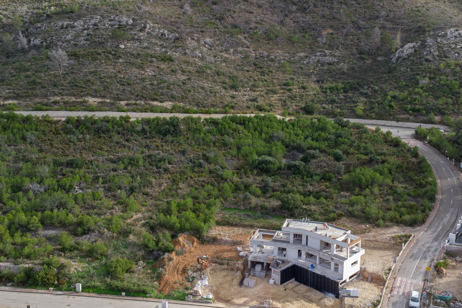 Vista aérea de una estructura en construcción en Monte Olímpico, Jávea, rodeada de vegetación autóctona y viales urbanizados con aceras y alumbrado.