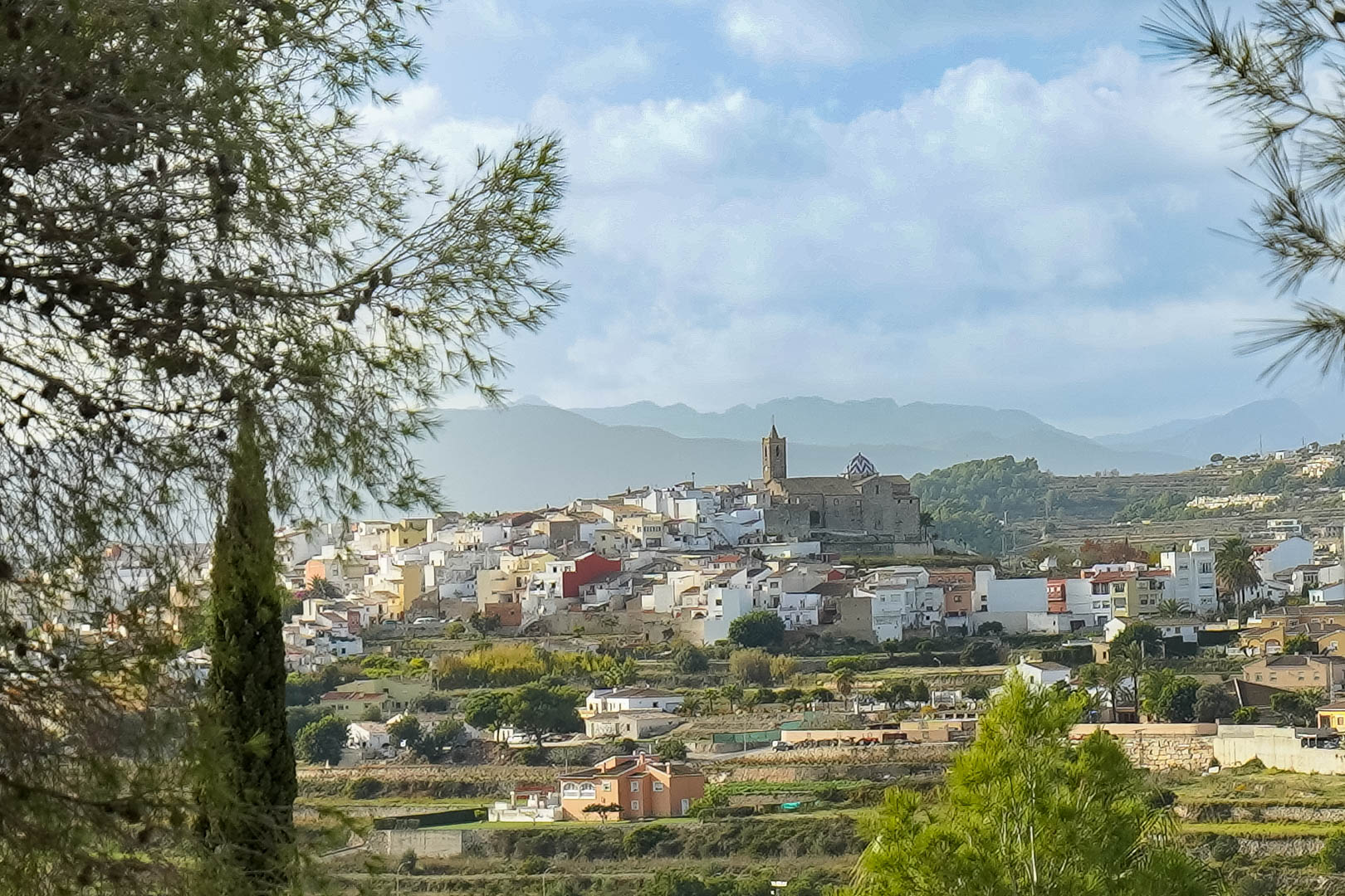Vistas panorámicas del casco histórico de Benitachell con su iglesia característica, enmarcadas por pinos y con la Sierra de Bernia al fondo.