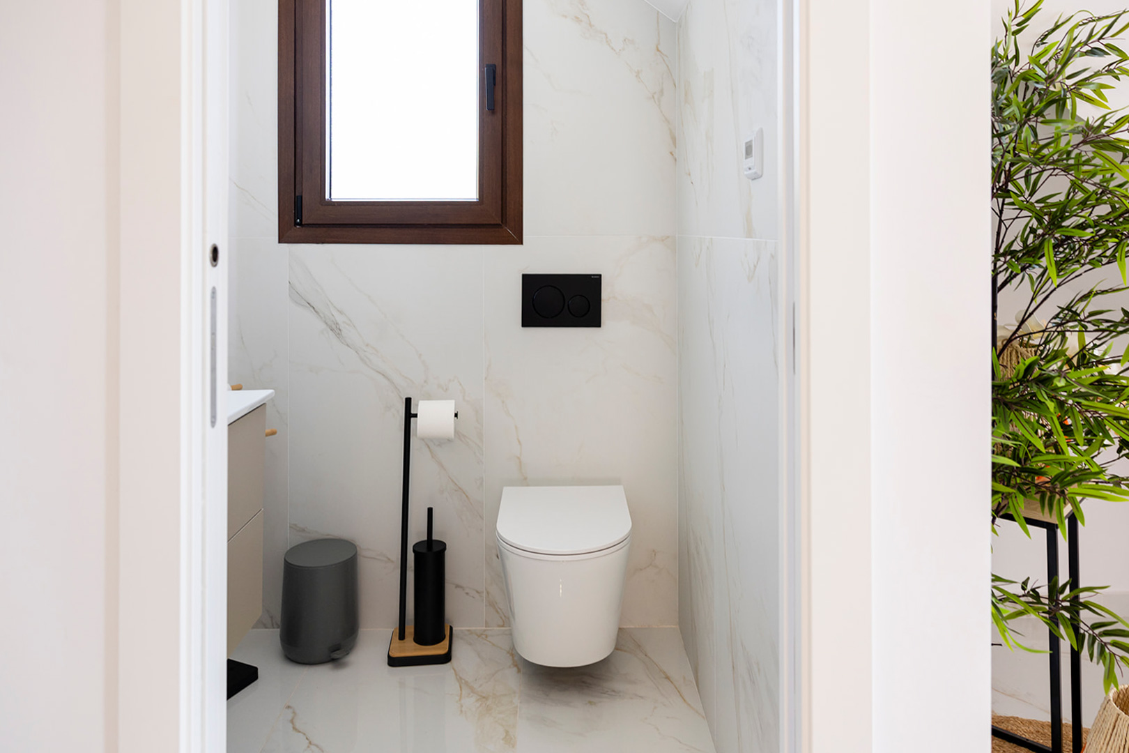 Aseo de cortesía con revestimiento porcelánico efecto mármol, inodoro suspendido y accesorios en negro mate bajo una ventana de marco oscuro.