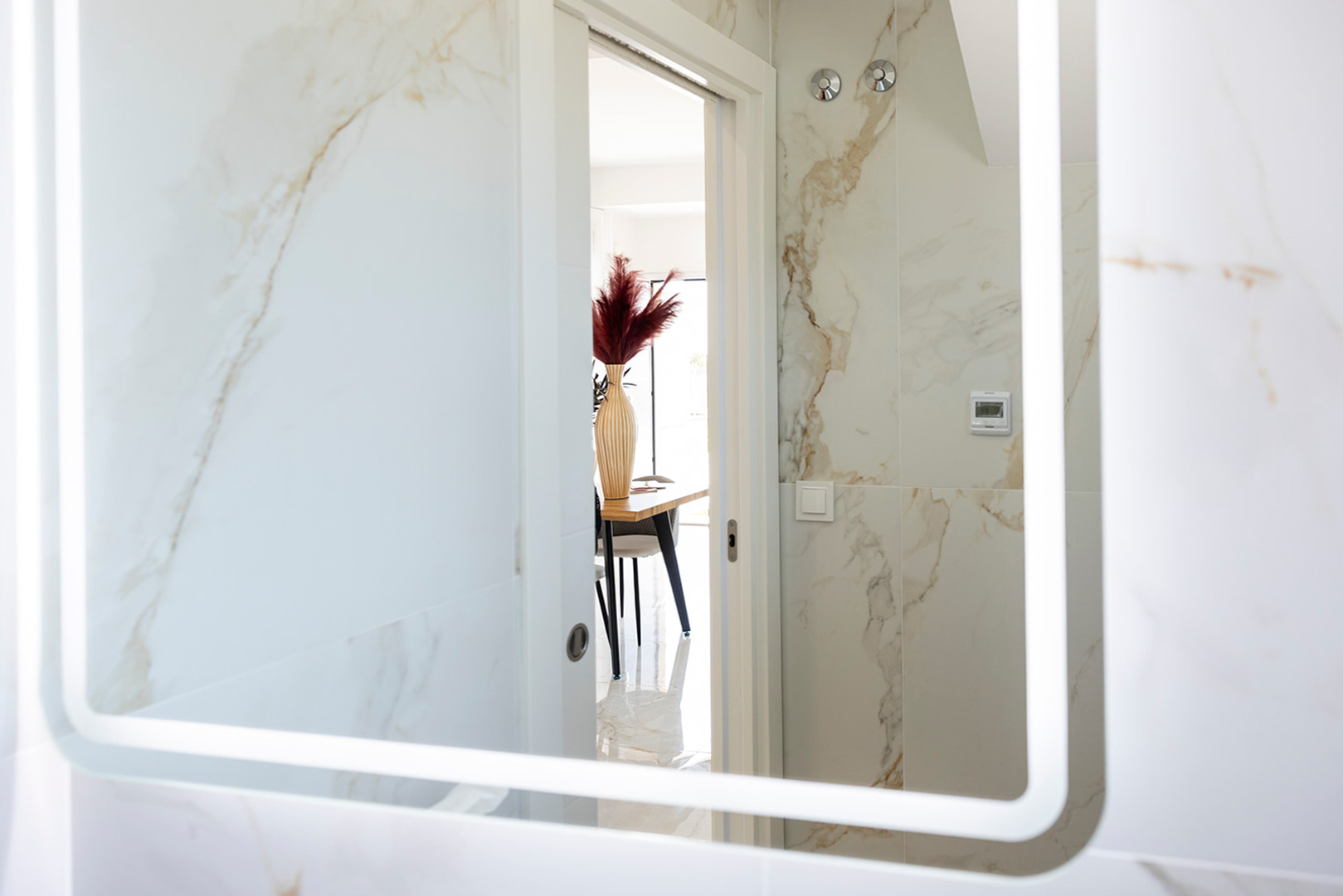 Baño con revestimiento cerámico efecto mármol y espejo retroiluminado, con vista a la zona de comedor a través de una puerta blanca.