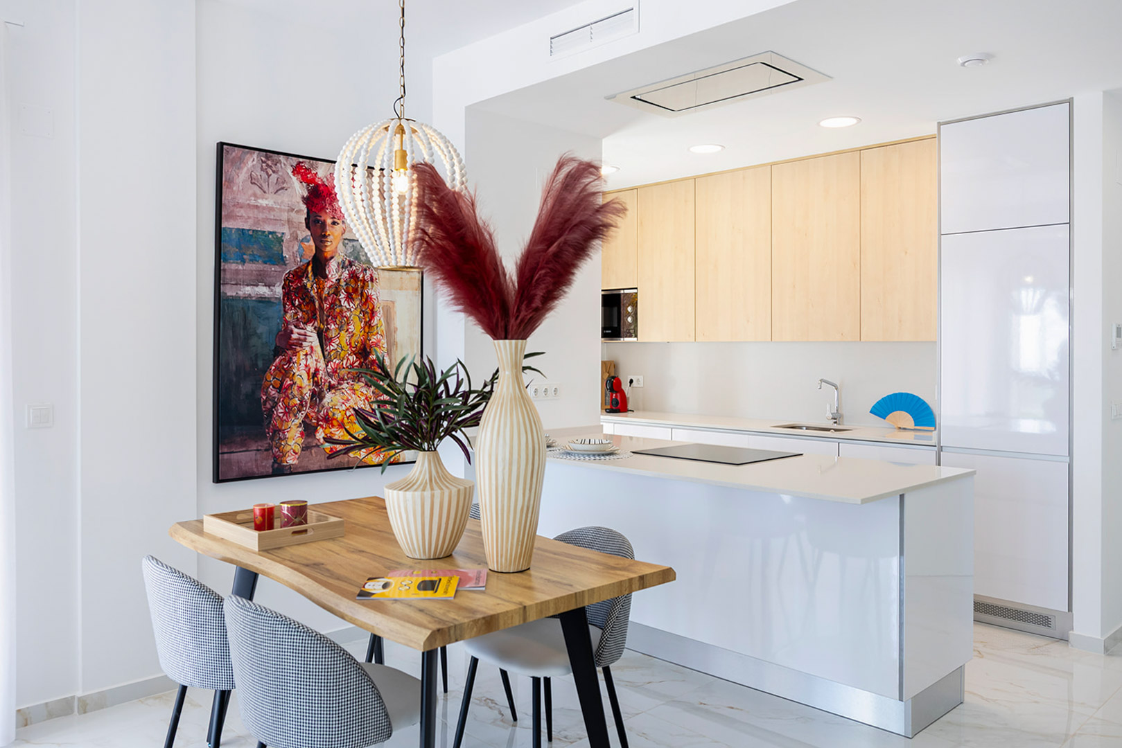 Cocina abierta con isla blanca y armarios de madera clara, integrada con un comedor de diseño que incluye mesa de madera, sillas tapizadas y lámpara de cuentas.