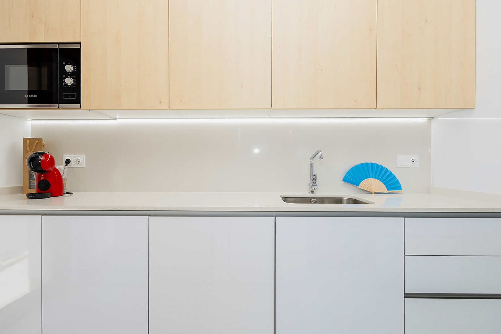 Cocina moderna con armarios superiores en madera clara e inferiores en blanco, encimera de cuarzo e iluminación LED integrada bajo los muebles.