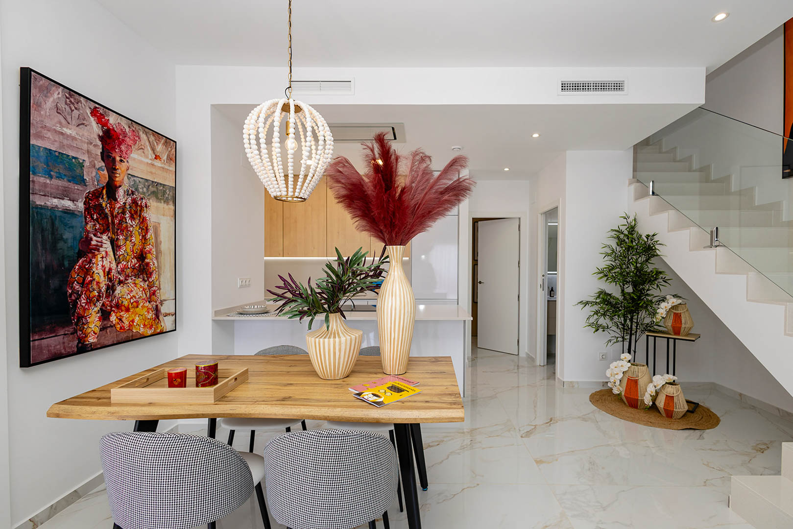 Comedor de concepto abierto con mesa de madera natural, lámpara colgante de cuentas y suelos de porcelánico efecto mármol junto a una escalera con barandilla de cristal.