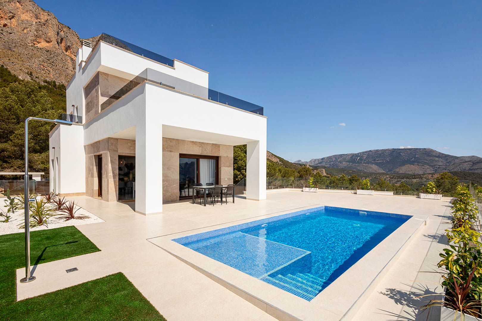 Exterior de villa moderna con fachada blanca y piedra, piscina privada de gresite azul, ducha de acero y terraza con vistas panorámicas a la montaña.