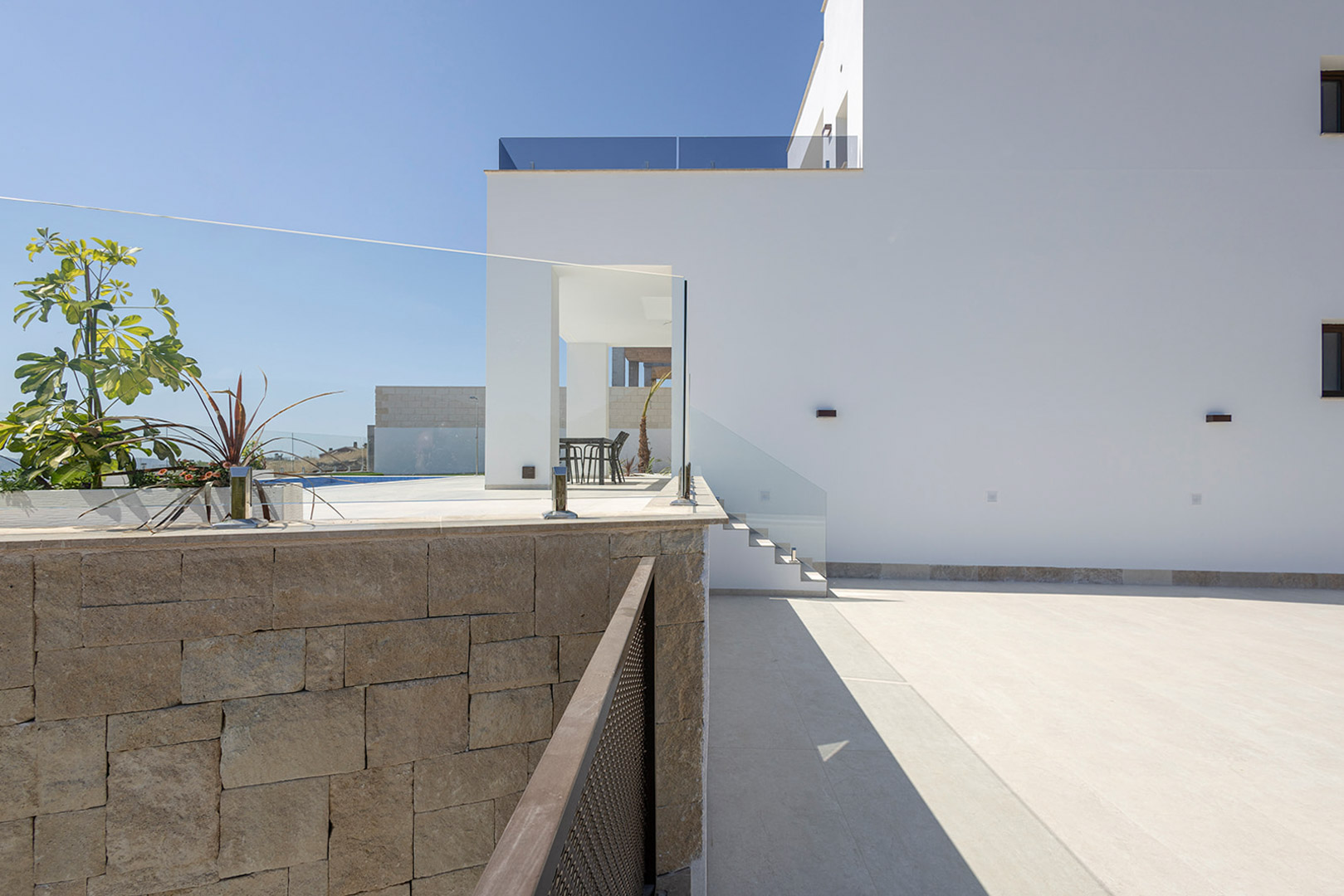 Fachada blanca de líneas rectas con muro de piedra natural, barandillas de cristal y zona exterior pavimentada con acceso a la terraza superior.