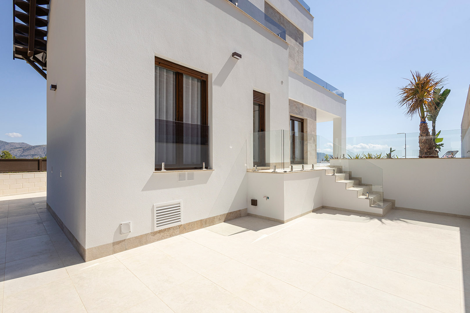 Fachada exterior de diseño minimalista con muros blancos, carpintería oscura y amplias terrazas pavimentadas con barandillas de cristal de seguridad.