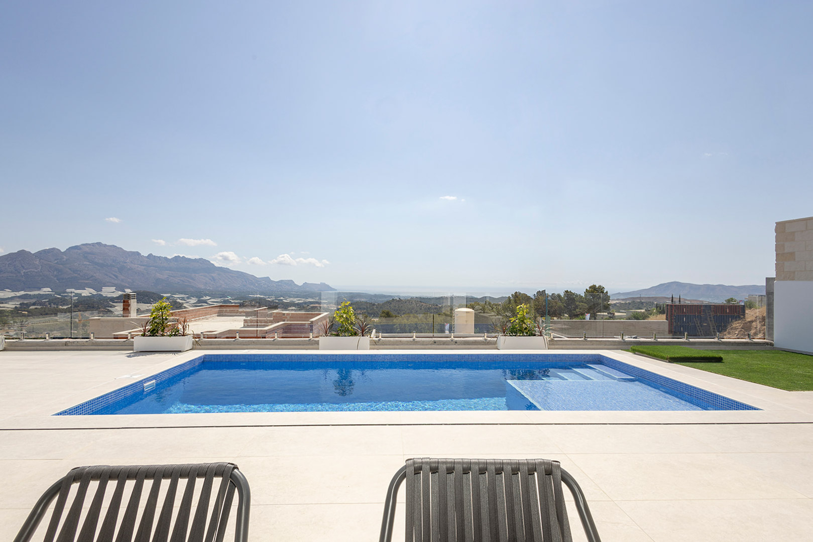 Piscina privada de gresite azul con escalera integrada, rodeada de una terraza de piedra clara y barandilla de cristal con vistas panorámicas al mar y la montaña.