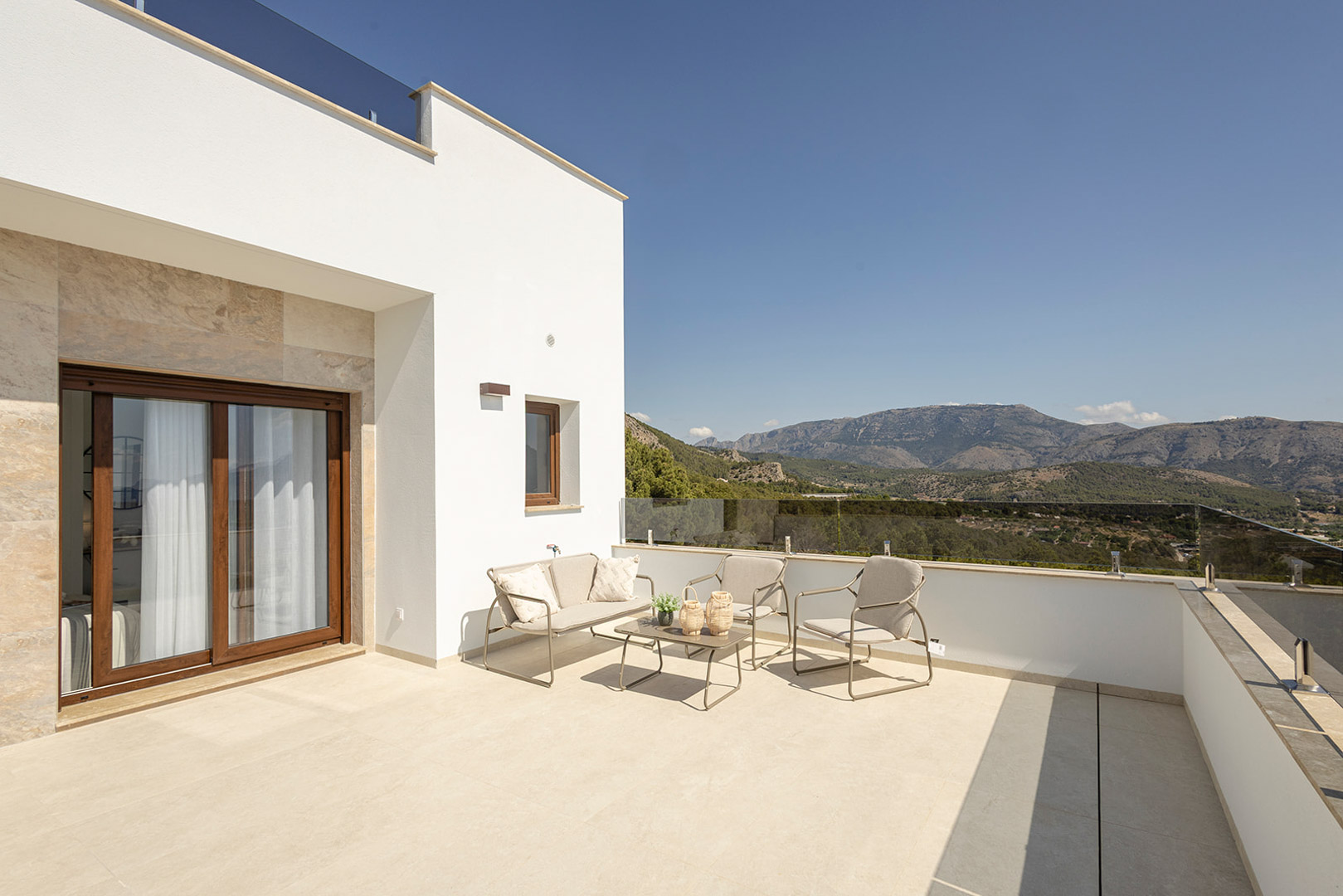 Terraza superior con barandilla de cristal, mobiliario de exterior y vistas panorámicas a las montañas. Fachada blanca con detalles en piedra y suelo cerámico claro.