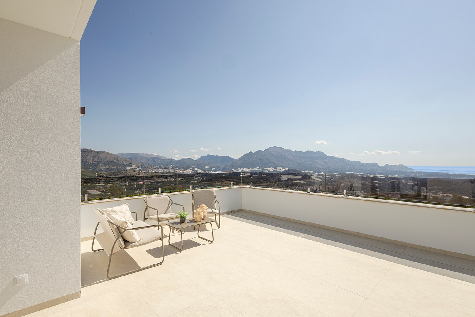 Terraza superior con barandilla de cristal, zona de estar exterior y vistas panorámicas a las montañas y al mar. Pavimento de gres porcelánico en tonos claros.