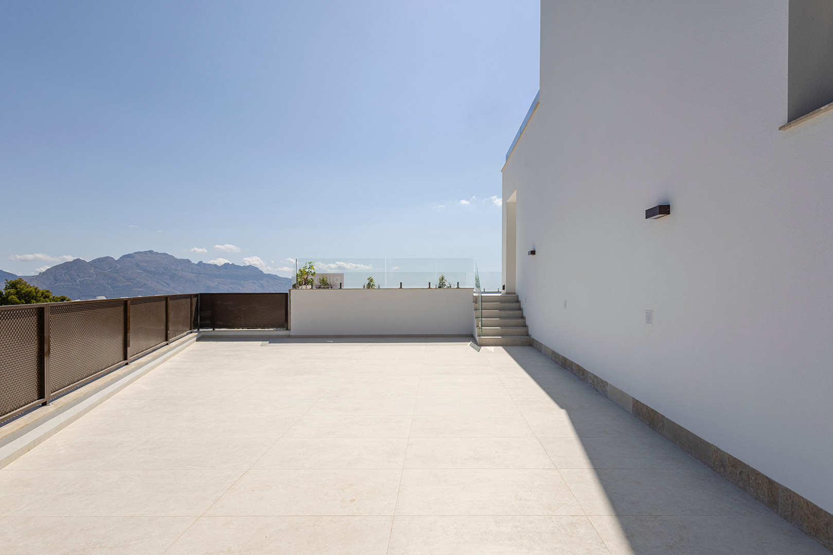Terraza superior con pavimento cerámico claro, barandillas de cristal y vistas despejadas a la montaña. Incluye iluminación exterior integrada en la fachada blanca.