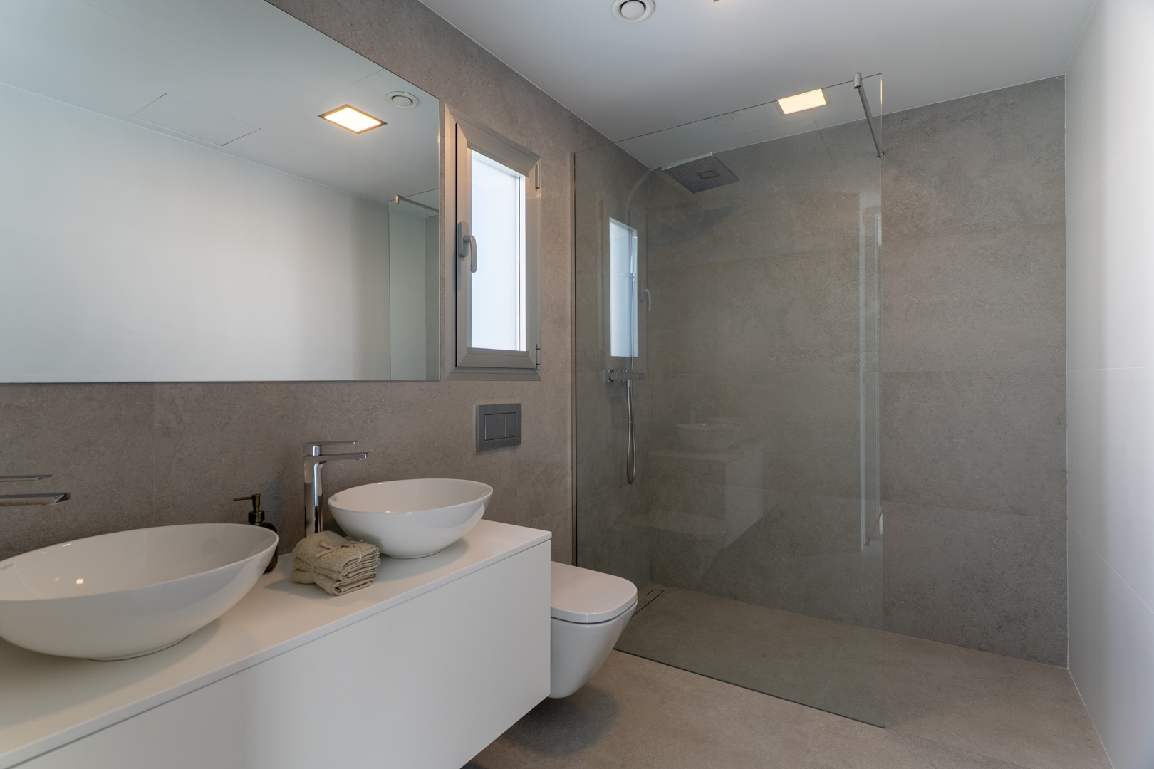 Baño moderno con revestimiento de gres porcelánico gris, lavabo doble sobre mueble suspendido blanco y ducha a ras de suelo con mampara de cristal fija.
