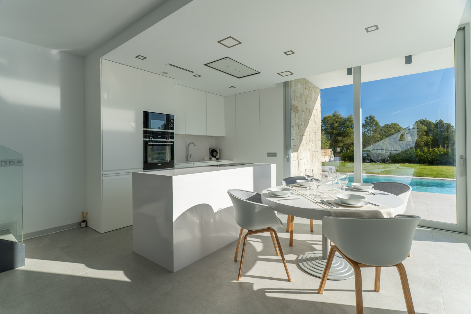 Cocina americana de diseño minimalista en blanco con isla central, integrada en un comedor luminoso con vistas directas a la piscina y el jardín privado.