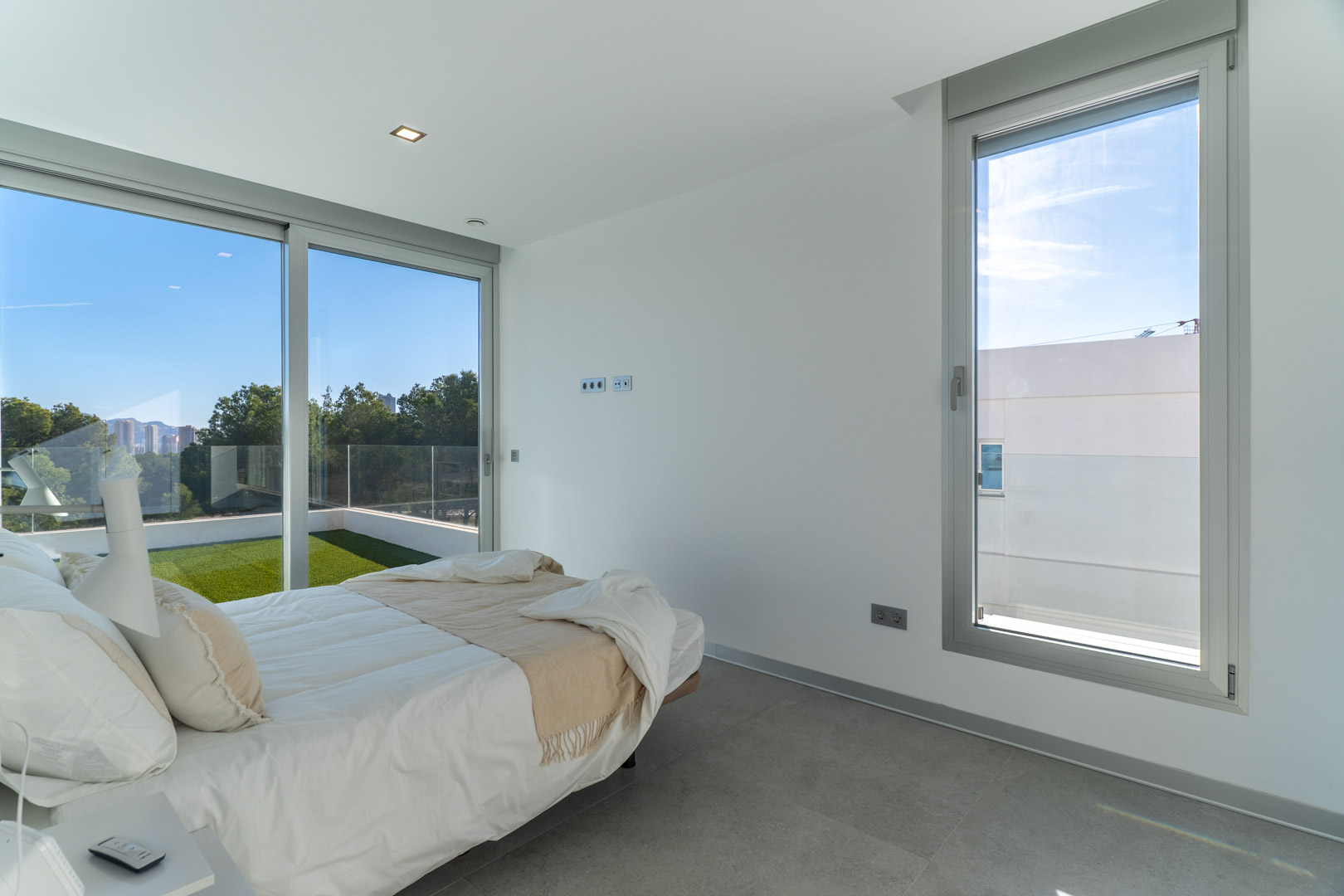 Dormitorio luminoso con suelos de gres gris, grandes ventanales de aluminio y acceso directo a una terraza privada con césped artificial y vistas despejadas.