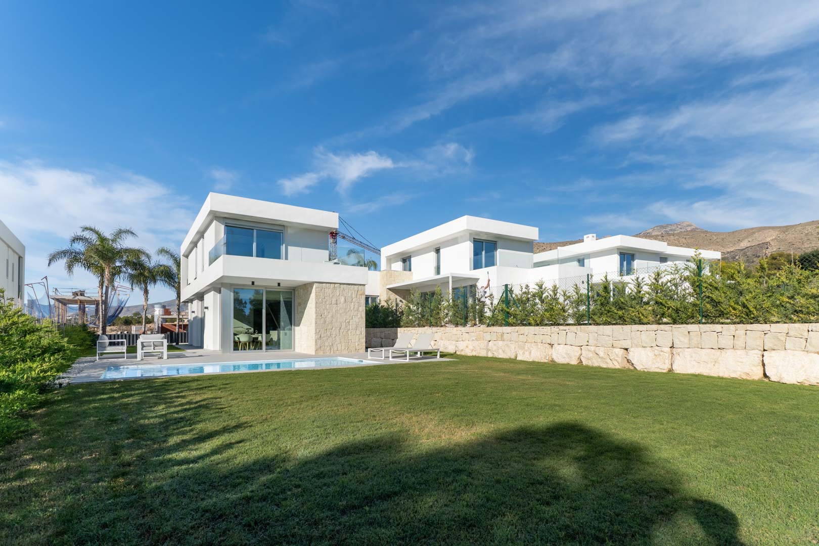 Exterior de villa moderna con fachada blanca y detalles en piedra, piscina privada, amplio jardín de césped y vistas a las montañas bajo un cielo despejado.