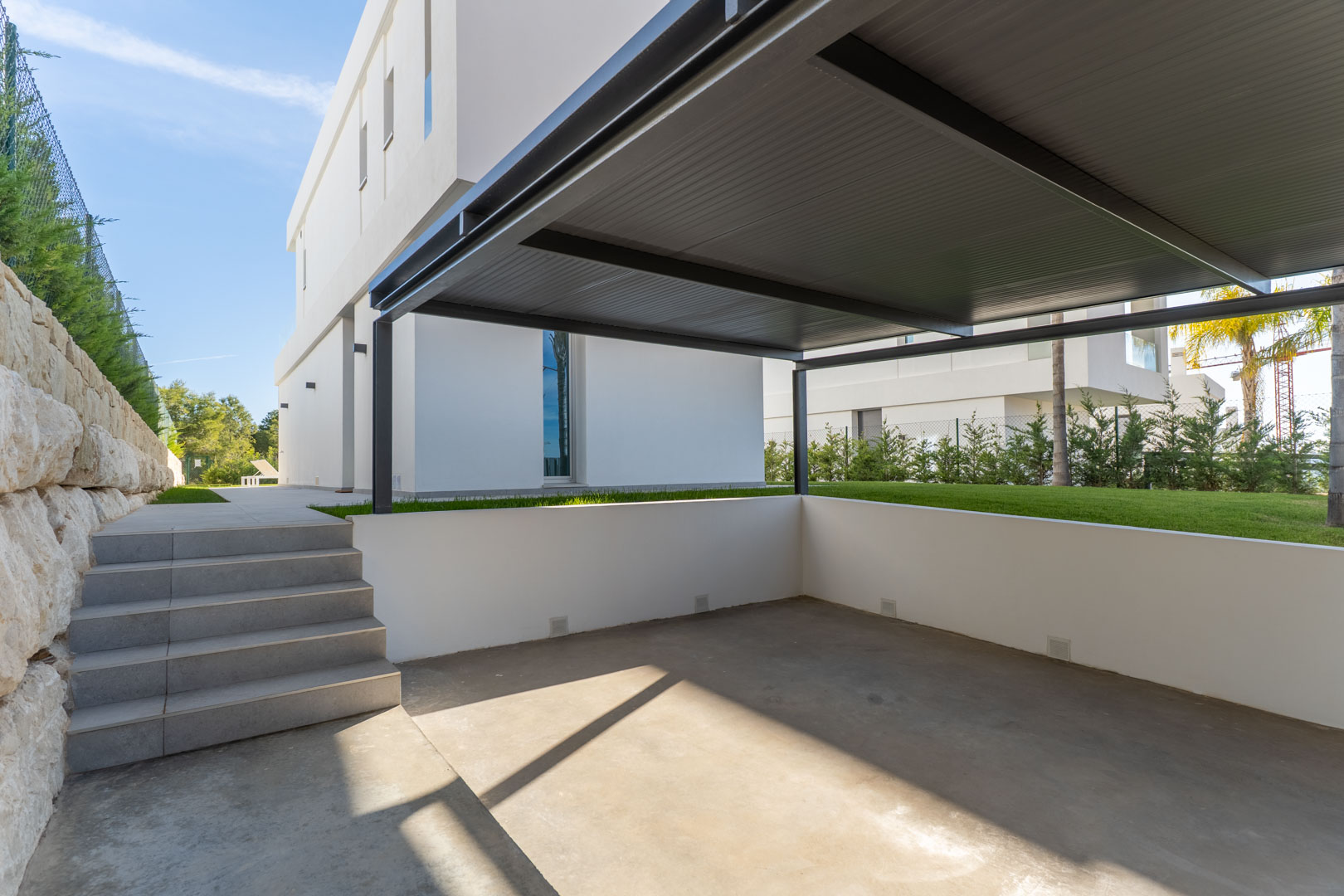 Zona de aparcamiento privado con pérgola metálica, suelo de hormigón y acceso mediante escaleras de gres a la vivienda y zona de jardín.