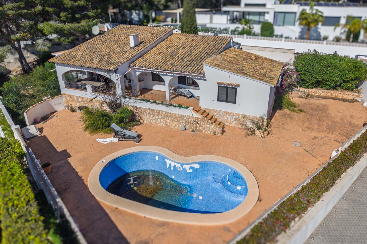Vista aérea de la villa con piscina de forma orgánica, amplias terrazas pavimentadas y porches arqueados. La fachada blanca destaca con muros de piedra natural y tejado tradicional.