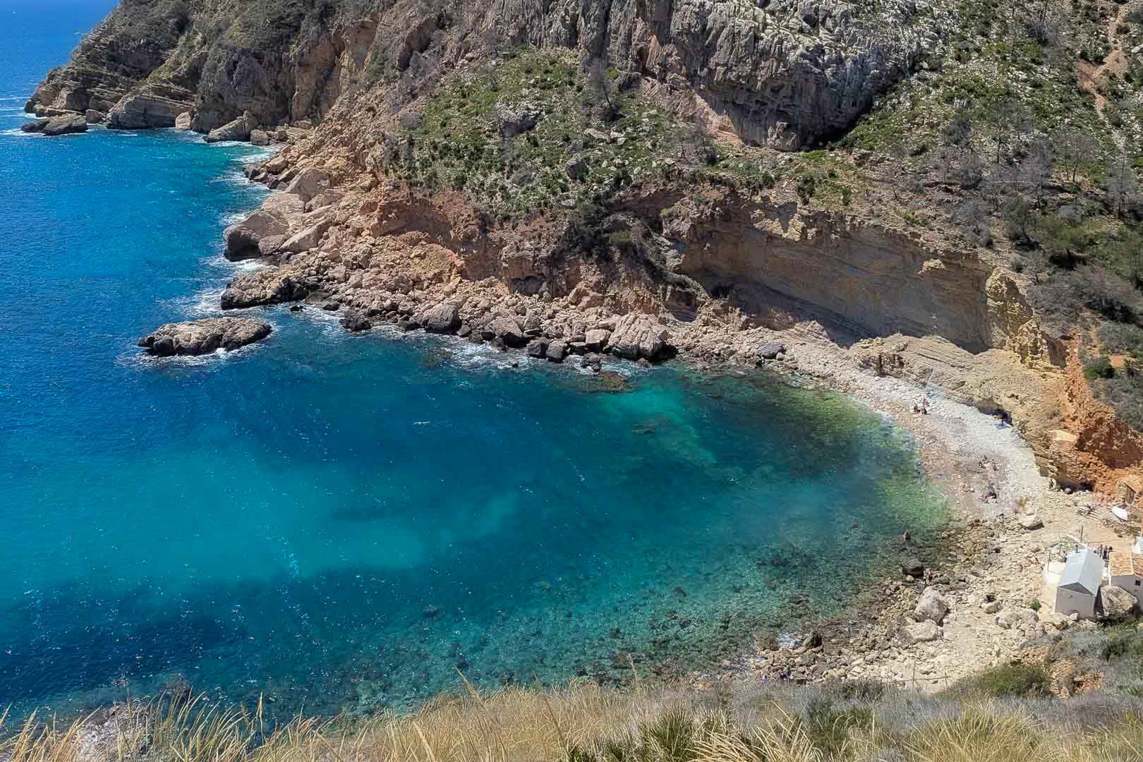 Vista aérea de una cala natural en Moraira con aguas cristalinas de color turquesa, acantilados escarpados y una pequeña playa de cantos rodados.