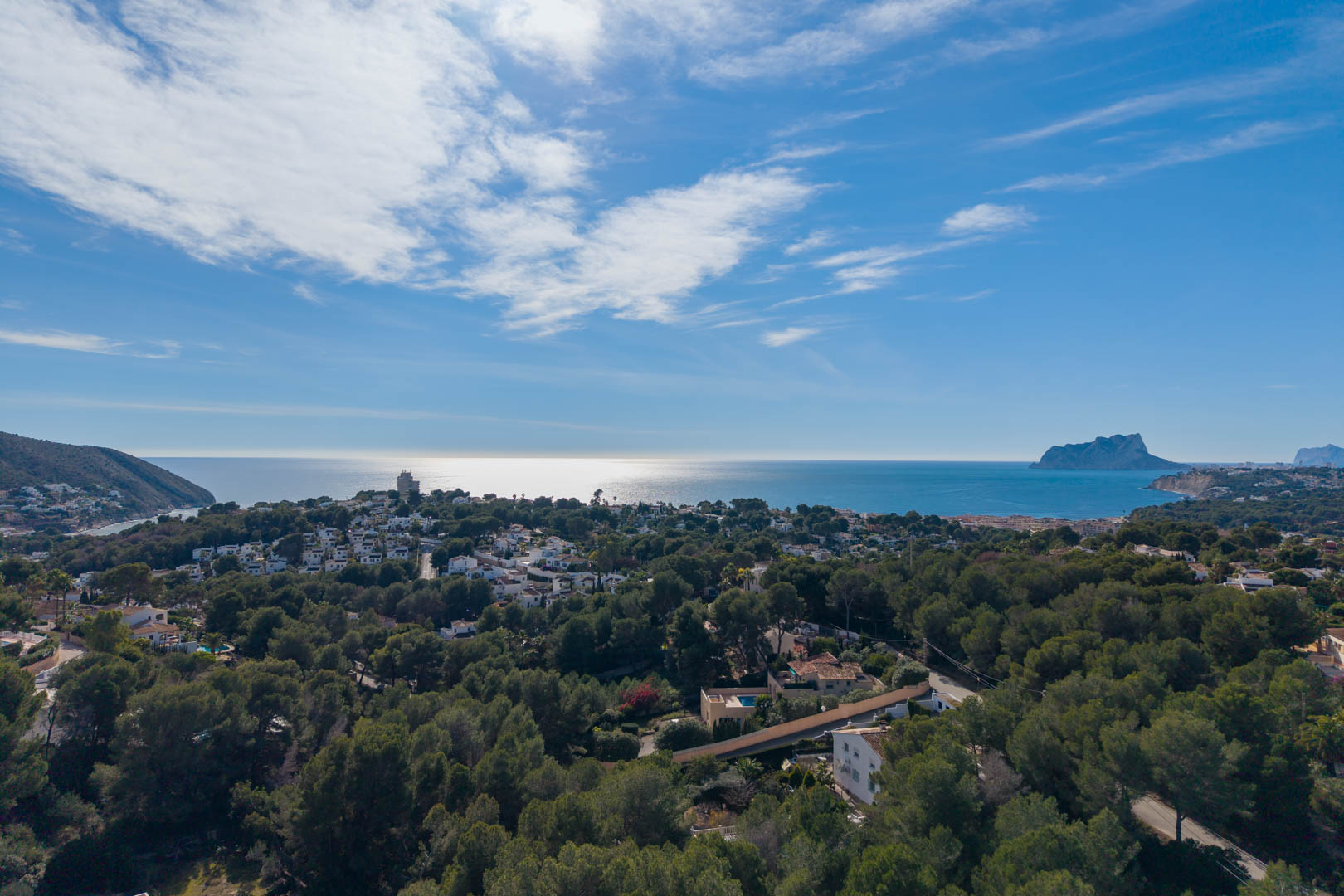 Vista aérea panorámica de la costa de Moraira, mostrando la densa vegetación de pinos, villas residenciales y el Peñón de Ifach sobre el horizonte del mar Mediterráneo.
