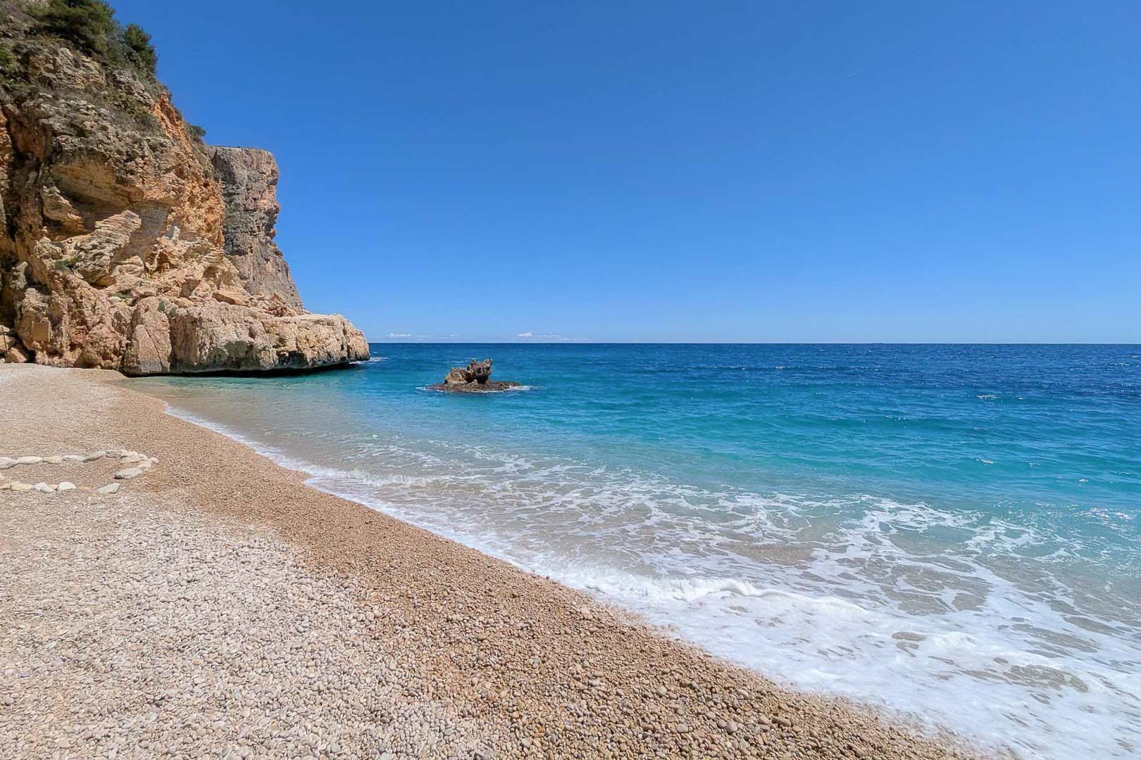 Vista de una cala de aguas cristalinas y playa de guijarros, flanqueada por acantilados de piedra caliza bajo un cielo despejado en la costa de Moraira.