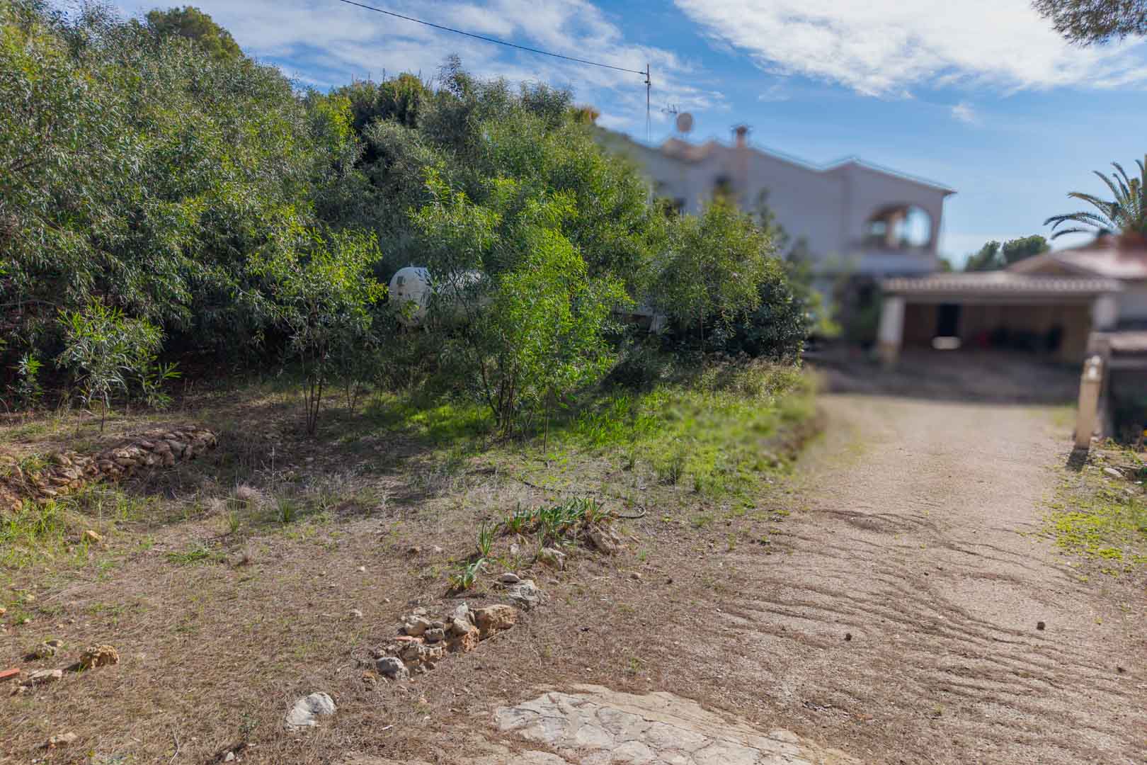 Vista del acceso a la parcela urbana, mostrando el terreno con vegetación autóctona y linderos de piedra natural junto a una vivienda existente.