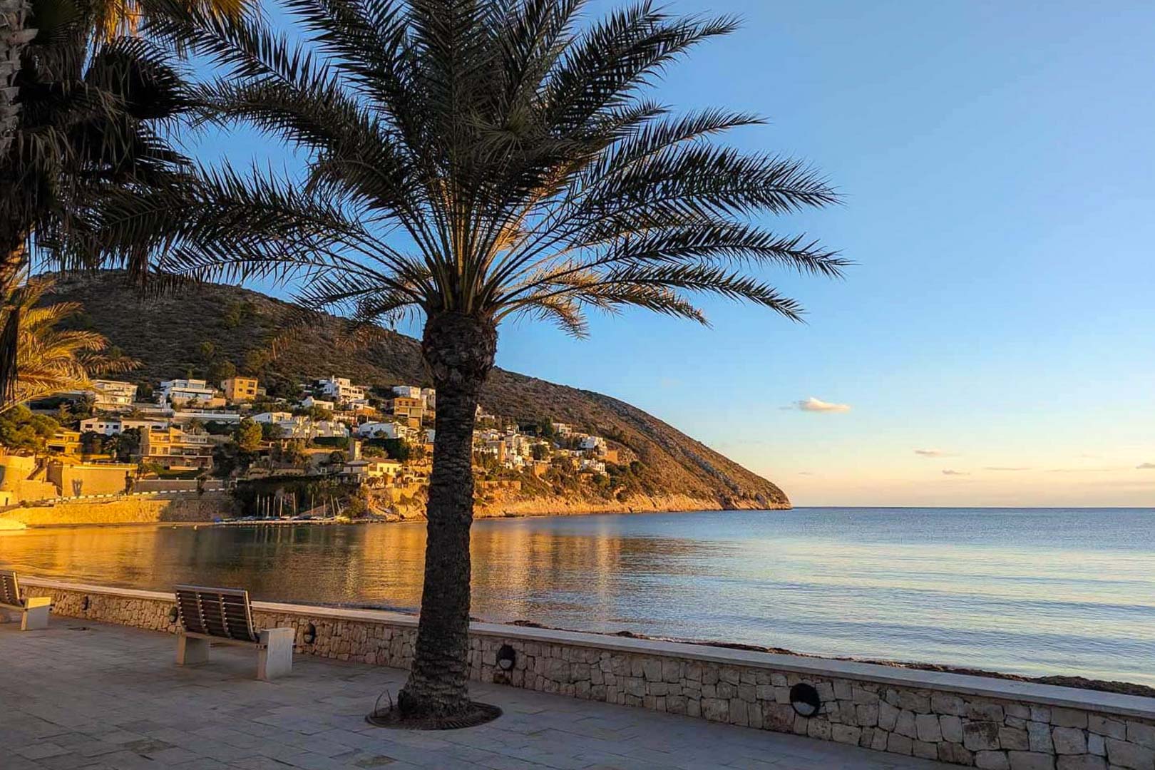Vista del paseo marítimo en la playa de El Portet, Moraira, con palmeras, muros de piedra natural y villas blancas integradas en la ladera costera.