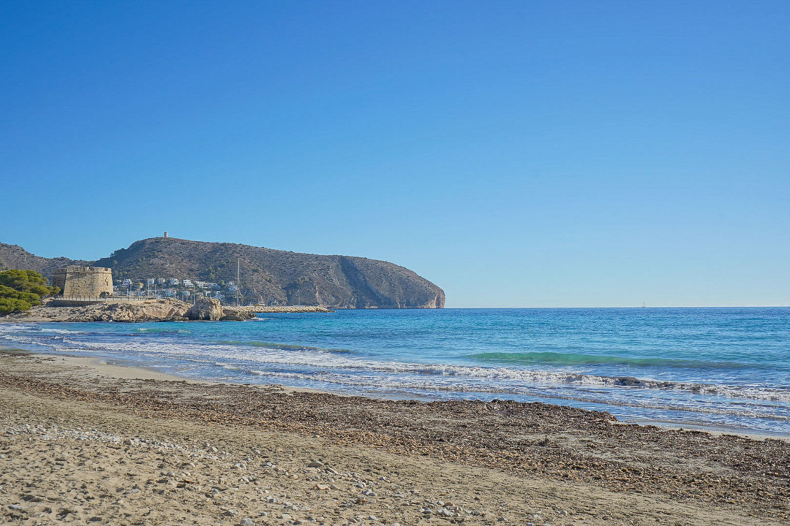 Vista panorámica de la playa de l'Ampolla en Moraira, mostrando el histórico torreón y el cabo de Cap d'Or bajo un cielo despejado y aguas cristalinas.