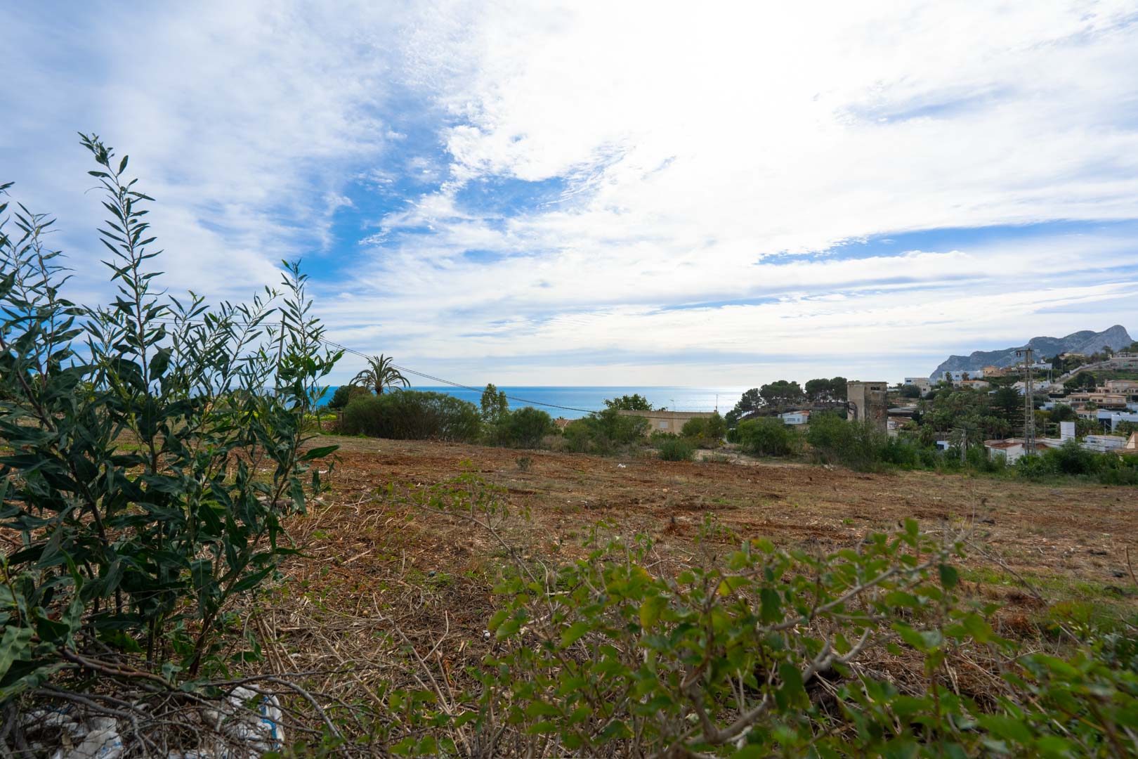 Parcela elevada con vistas panorámicas al mar Mediterráneo y las montañas, rodeada de vegetación autóctona y arquitectura residencial costera.