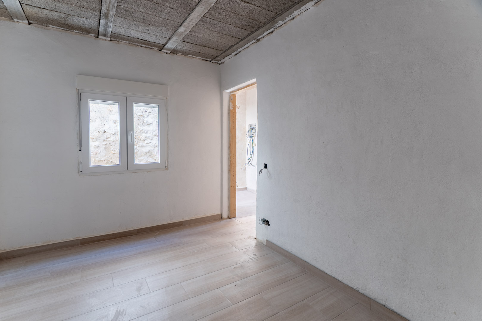 Habitación en reforma con suelos cerámicos de imitación madera, paredes blancas y ventana de PVC. Se aprecia el techo estructural y la preinstalación eléctrica.