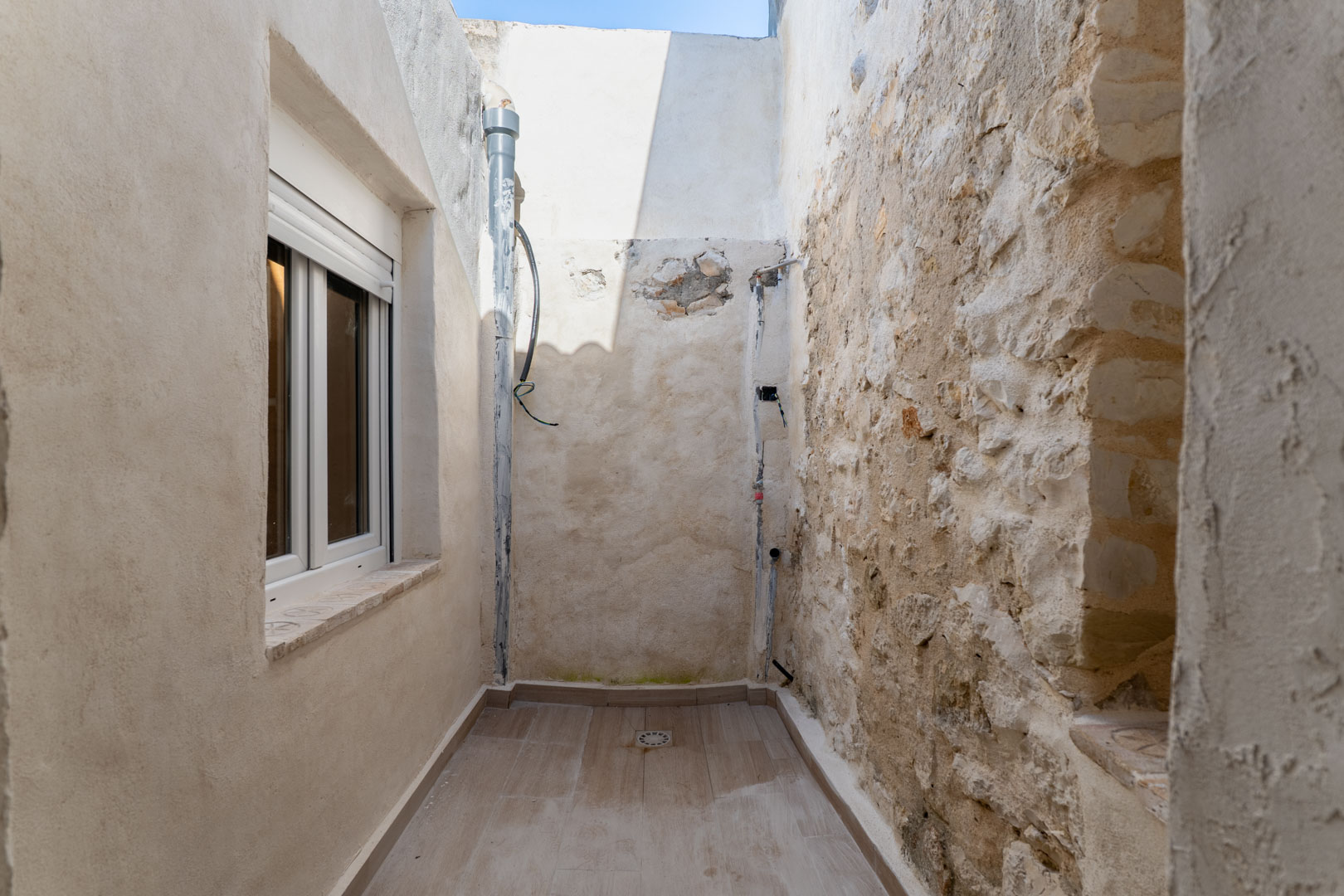 Patio interior con suelo cerámico y desagüe, que combina un muro original de piedra seca con una fachada reformada y ventana de PVC blanco.