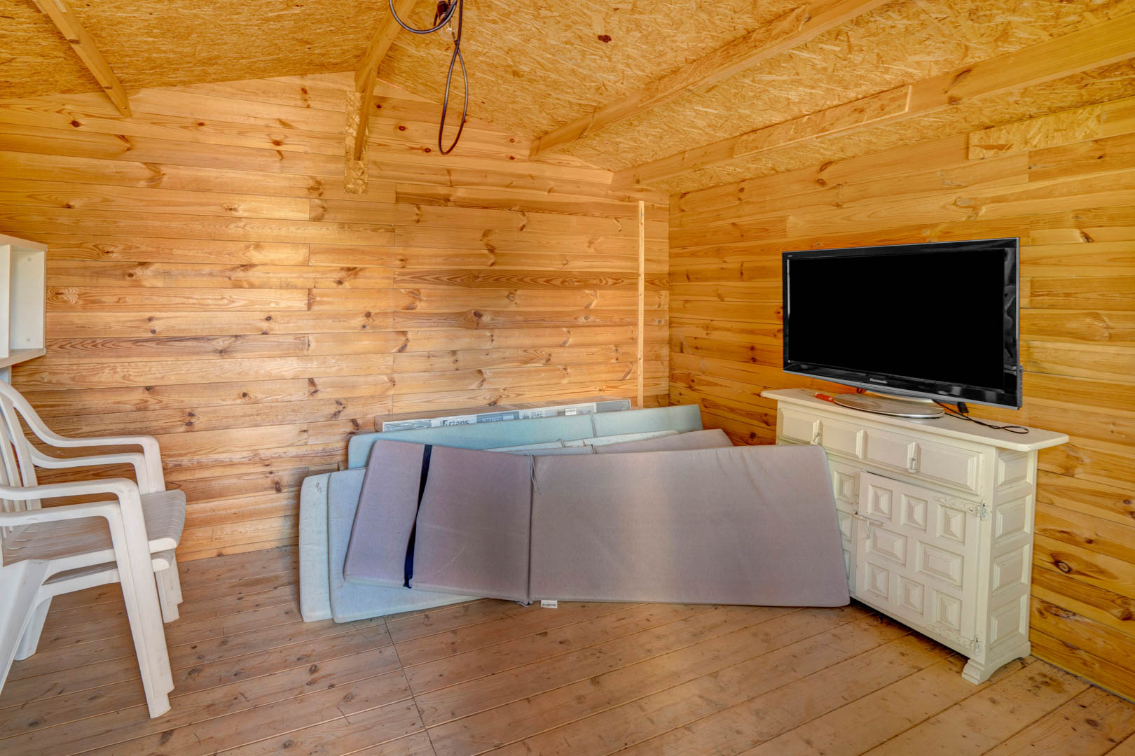 Caseta de madera o trastero con paredes y suelos de pino, equipada con mobiliario auxiliar, televisión y espacio de almacenamiento para cojines de exterior.