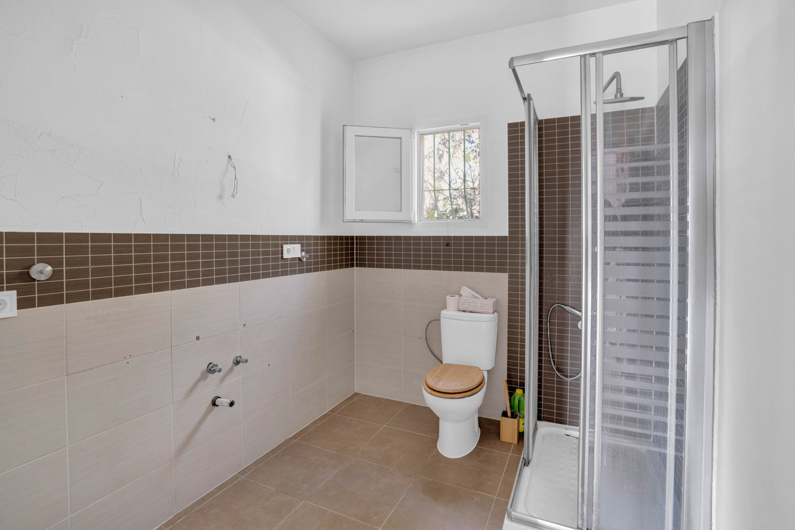 Cuarto de baño con plato de ducha y mampara, inodoro con tapa de madera y paredes alicatadas en tonos tierra con cenefa de mosaico marrón. Incluye ventana para ventilación natural.