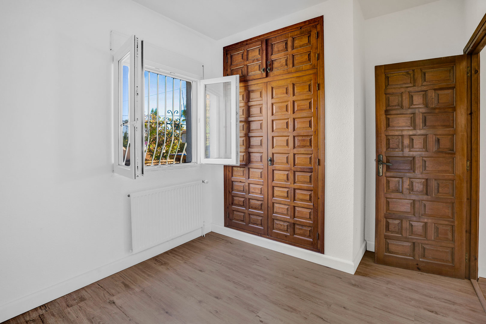 Dormitorio con armario empotrado de madera tallada, suelos laminados, radiador de calefacción central y ventana con luz natural y rejas de seguridad.