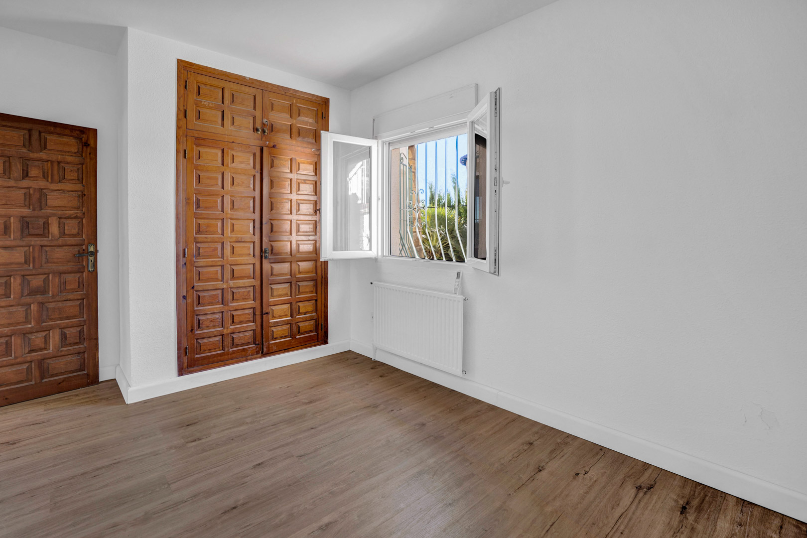 Dormitorio luminoso con armario empotrado de madera tallada, suelos laminados, calefacción por radiador y ventana de PVC con vistas al jardín.