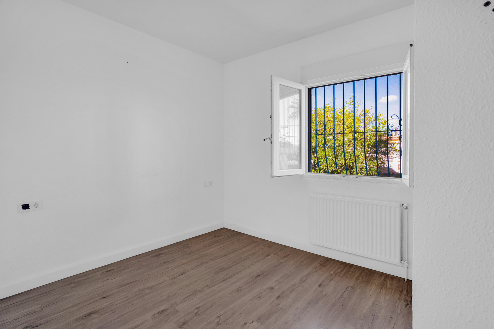 Habitación luminosa con suelos de tarima laminada, paredes blancas y ventana con rejas decorativas que ofrece vistas al jardín y calefacción por radiador.