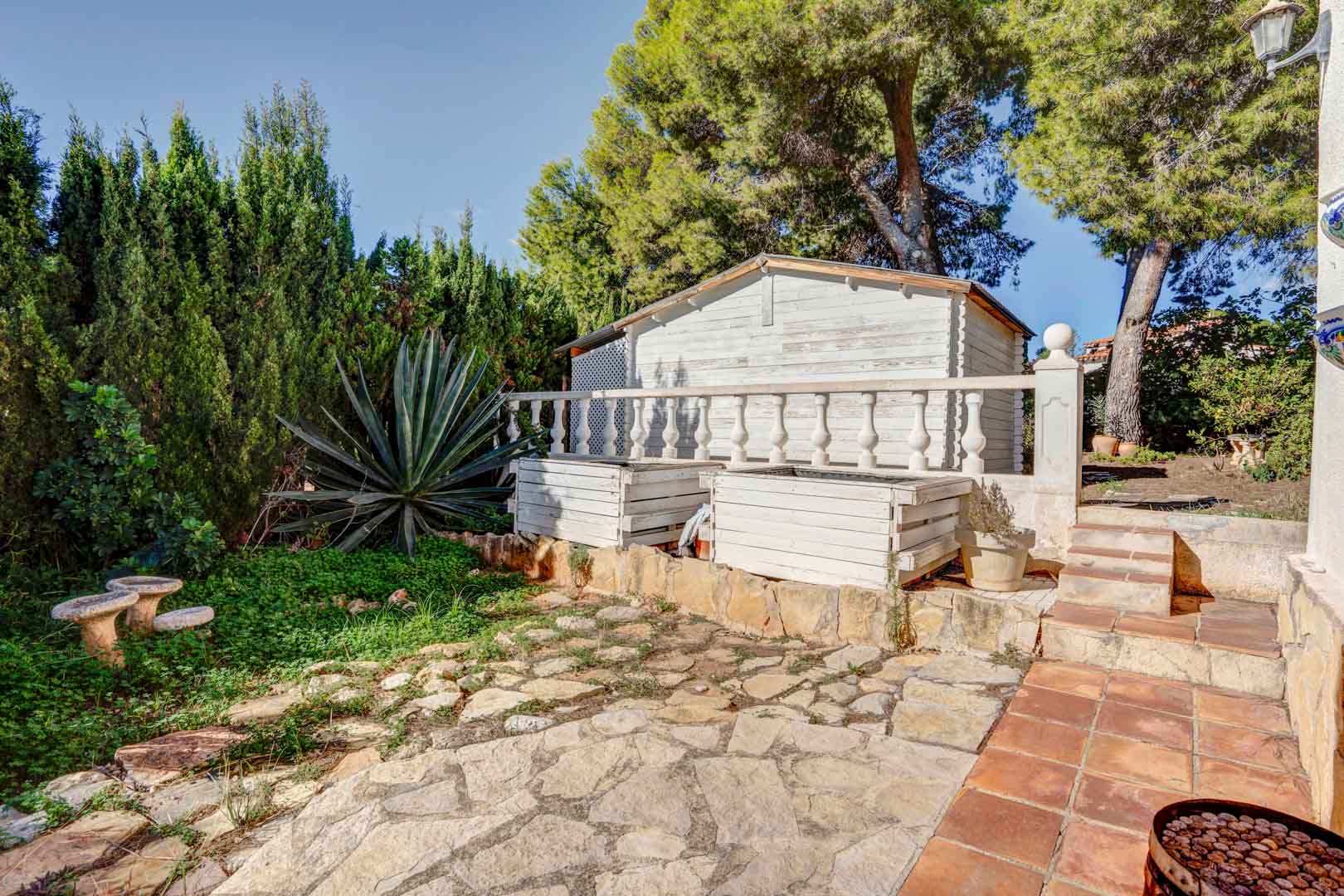 Jardín con pavimentación de piedra irregular, caseta de madera blanca y vegetación mediterránea madura con pinos y agaves bajo luz natural.