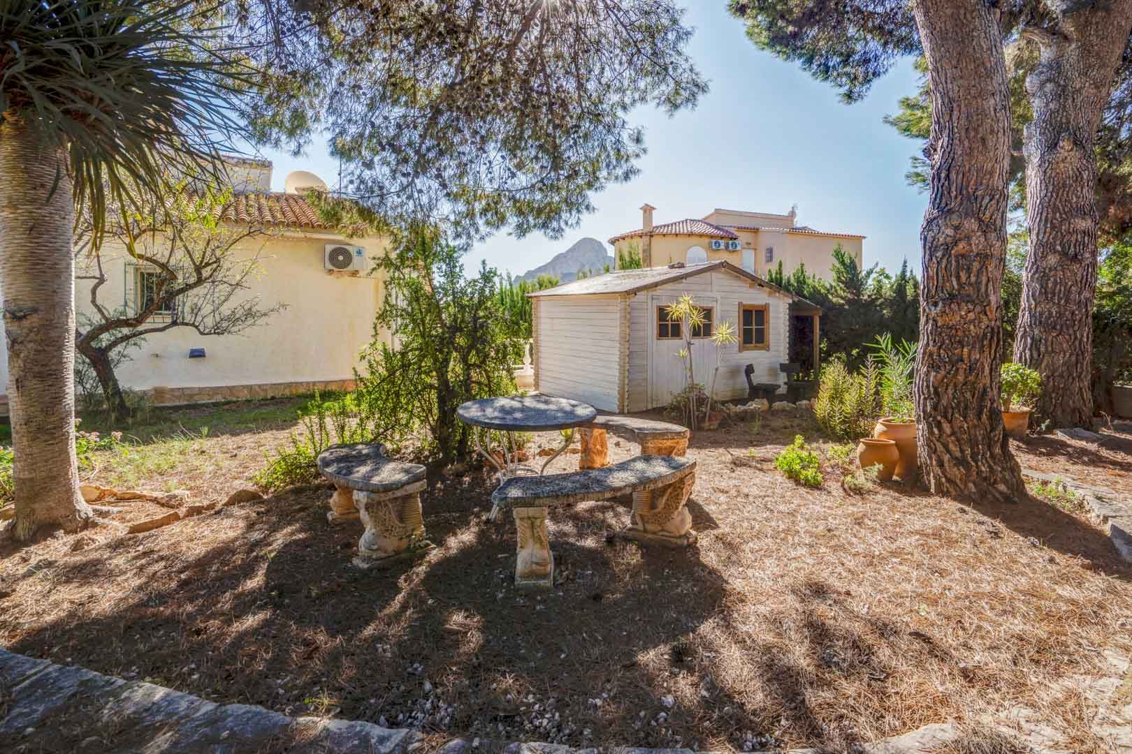 Jardín mediterráneo con zona de descanso bajo pinos, mesa y bancos de piedra, caseta de madera blanca y vistas lejanas al Peñón de Ifach.