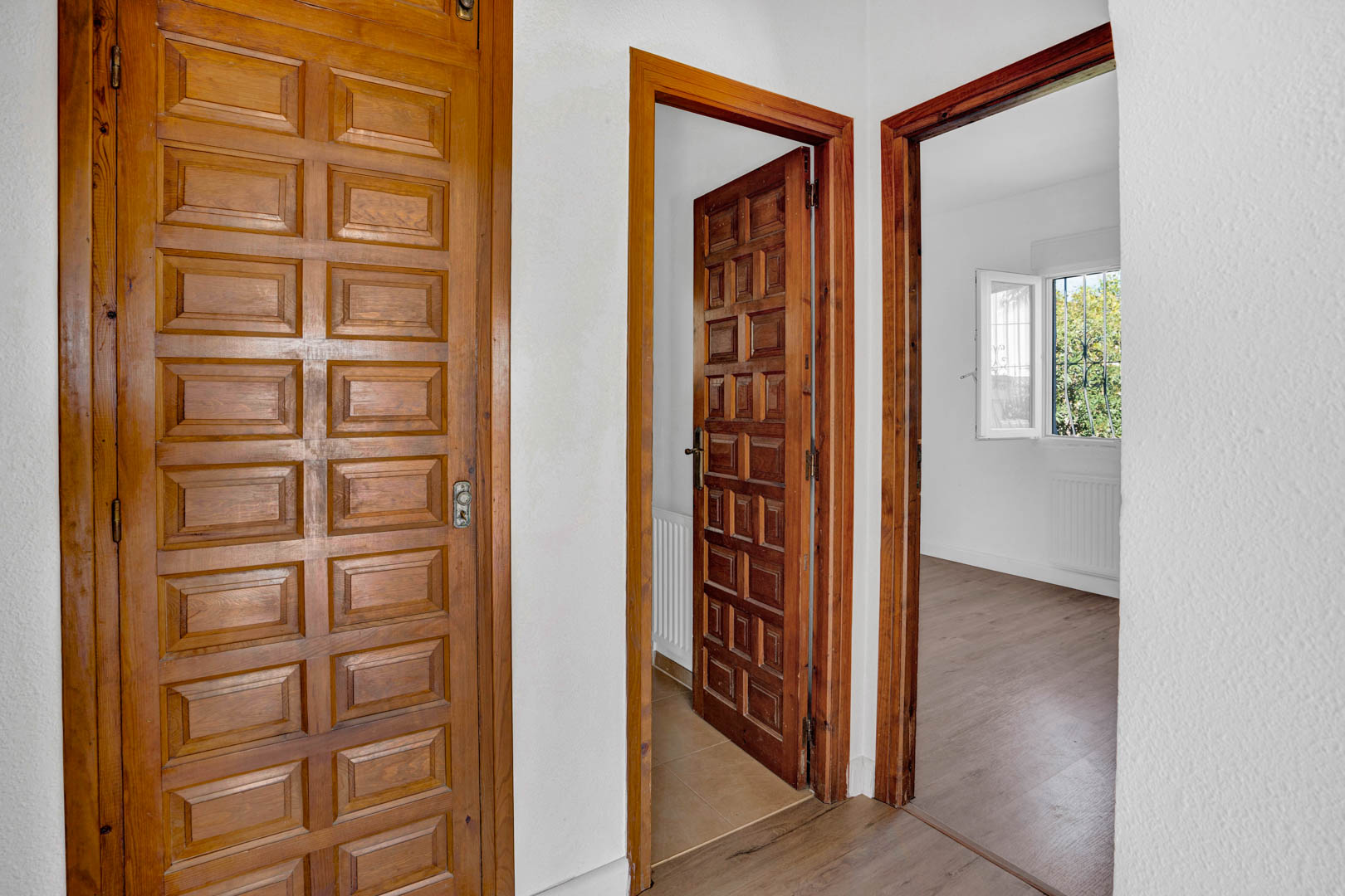 Pasillo distribuidor con puertas de madera maciza paneladas, paredes blancas y suelos laminados que conectan con dormitorios luminosos y exteriores.