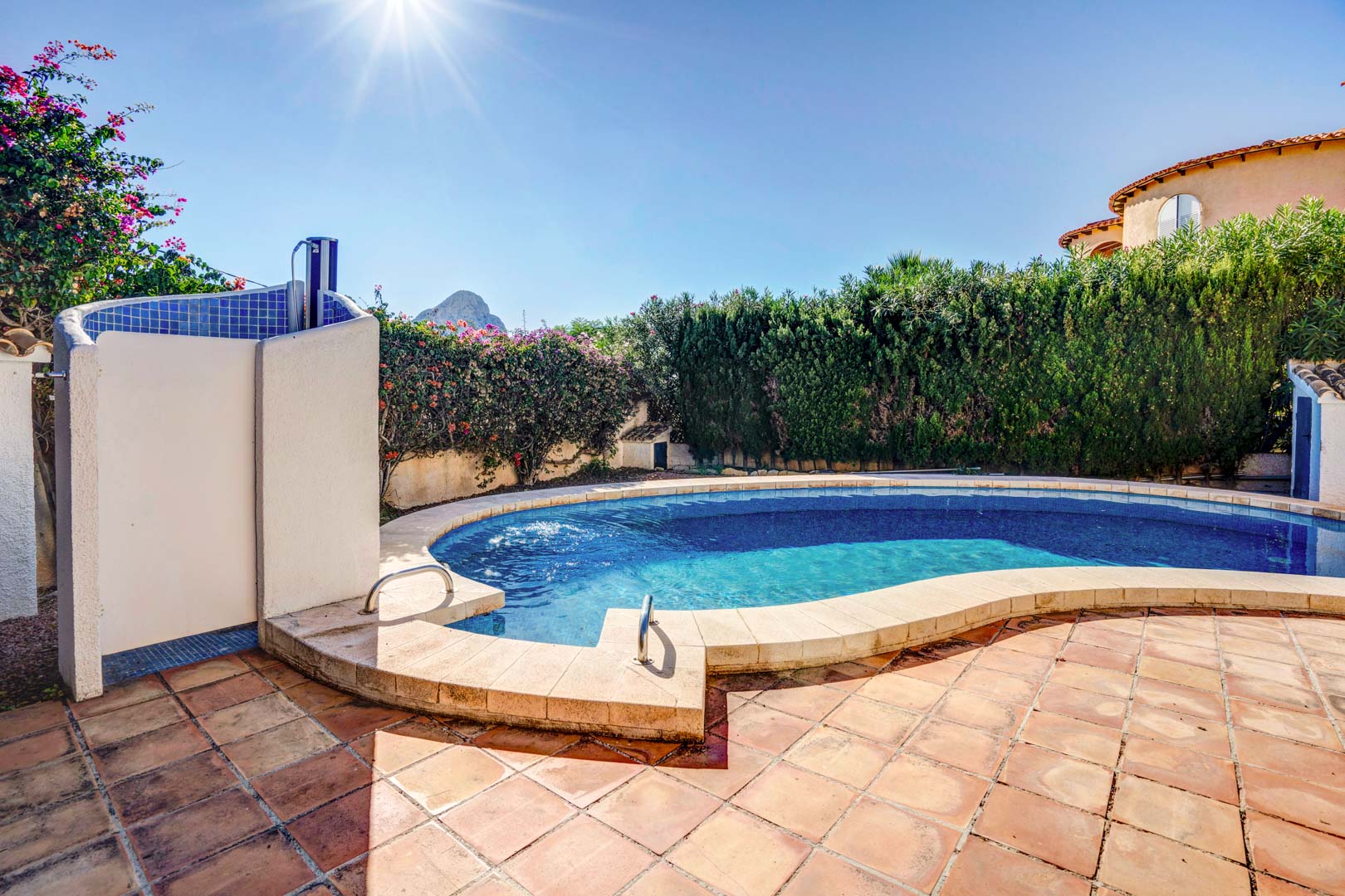 Piscina privada de forma curva rodeada de una terraza de terracota, con ducha exterior y vistas al Peñón de Ifach tras setos que garantizan total privacidad.