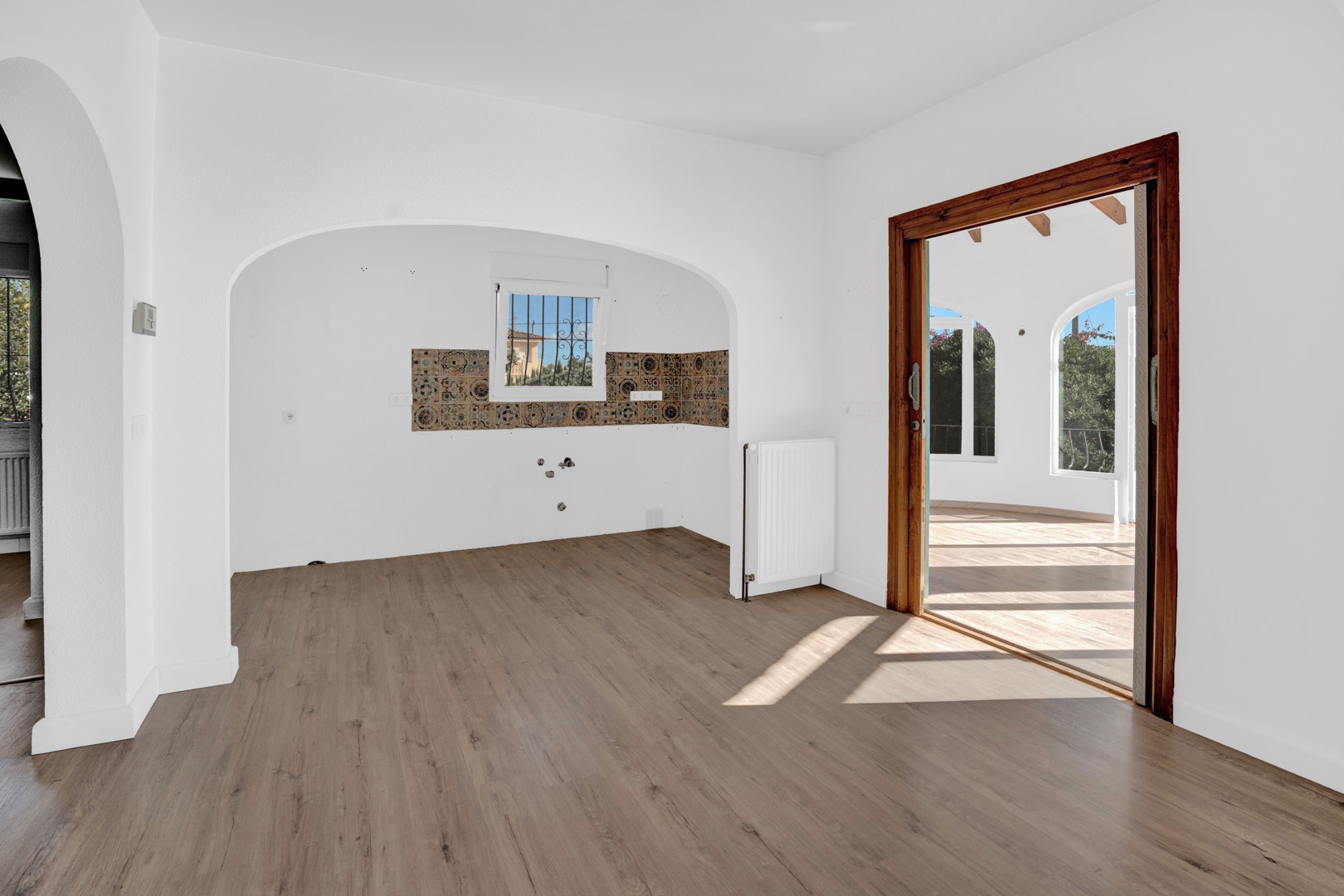 Salón comedor con suelos laminados, radiador y arco hacia la cocina con azulejos decorativos. Una puerta corredera de madera da acceso a la terraza acristalada.