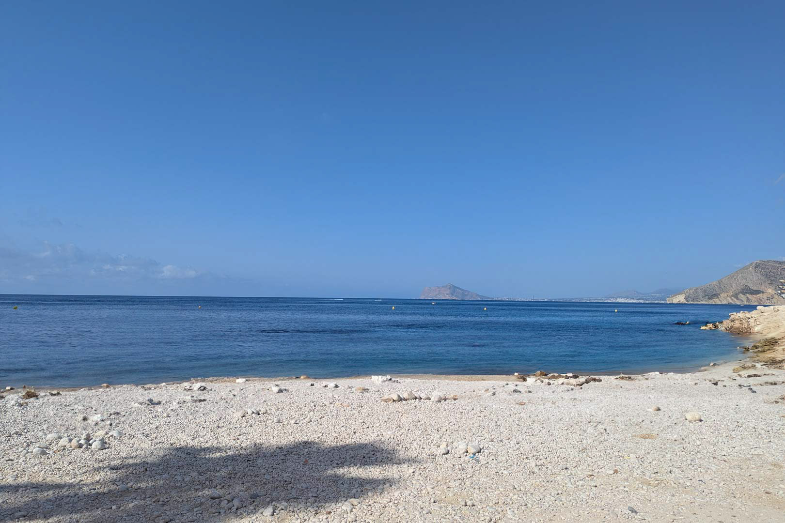 Vista de una cala de cantos blancos y aguas tranquilas en Calpe, con el perfil de la costa mediterránea y montañas al fondo bajo un cielo despejado.