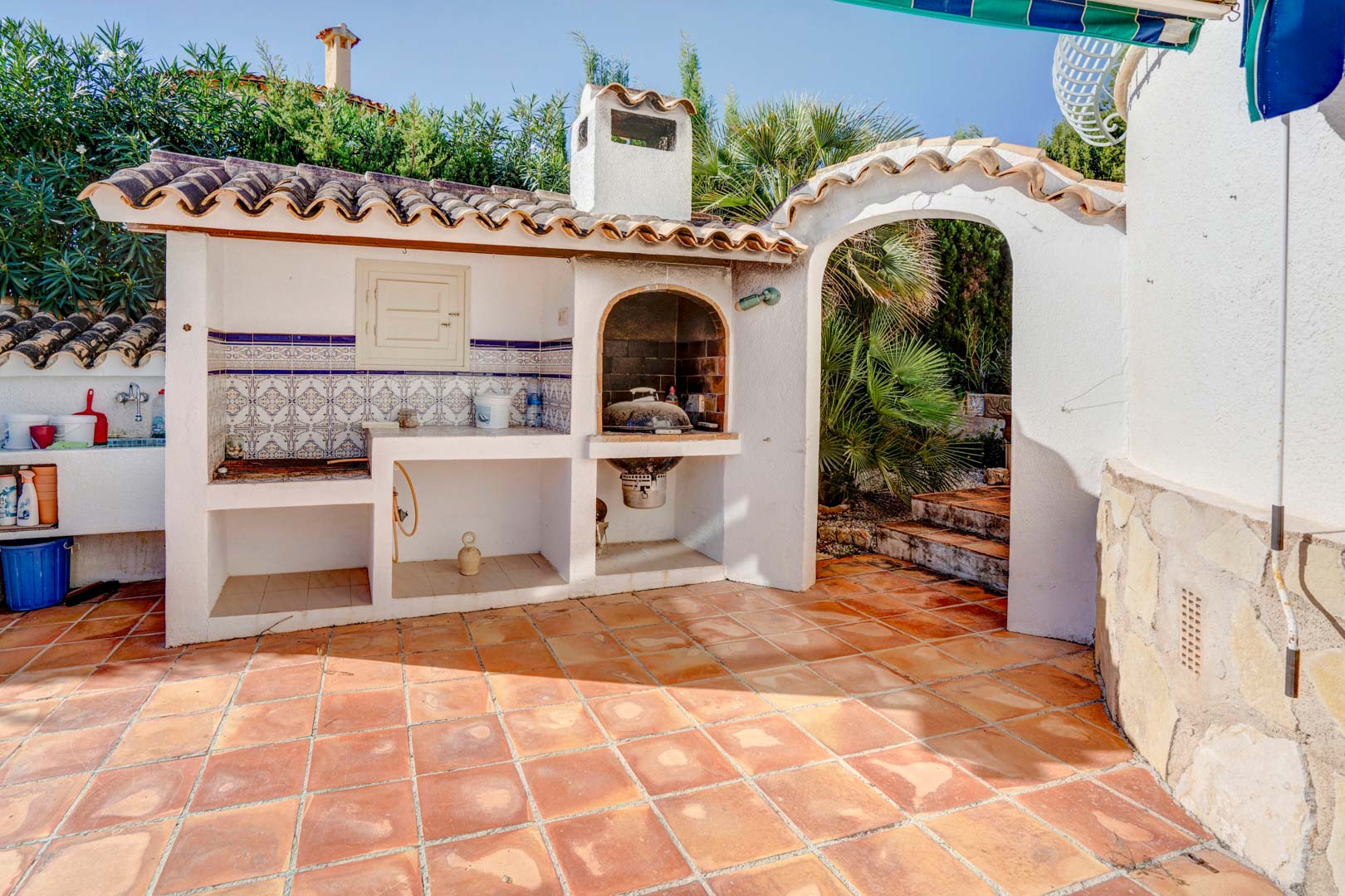 Zona de barbacoa de obra con chimenea, encimera de azulejos tradicionales y suelo de terracota, situada junto a un arco de acceso al jardín mediterráneo.