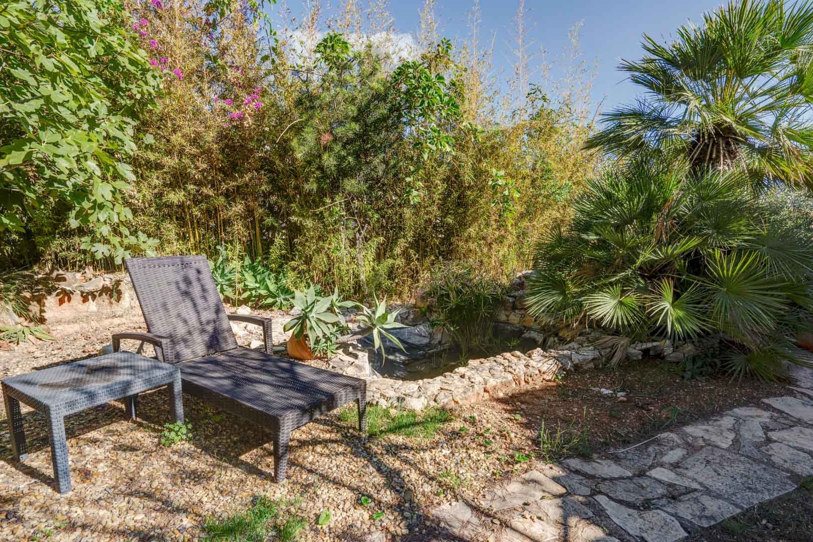 Zona de descanso en el jardín con tumbona de ratán sintético junto a un estanque de piedra natural, rodeado de vegetación mediterránea, palmeras y bambú.